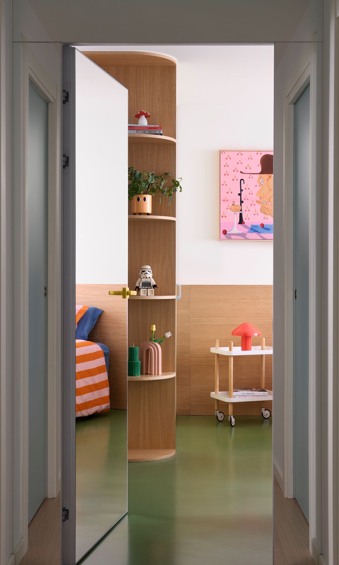 Entrada a habitación con puerta de espejo, estantería curva en una esquina, cuadro en la pared, mesita de noche con ruedas, suelo verde 