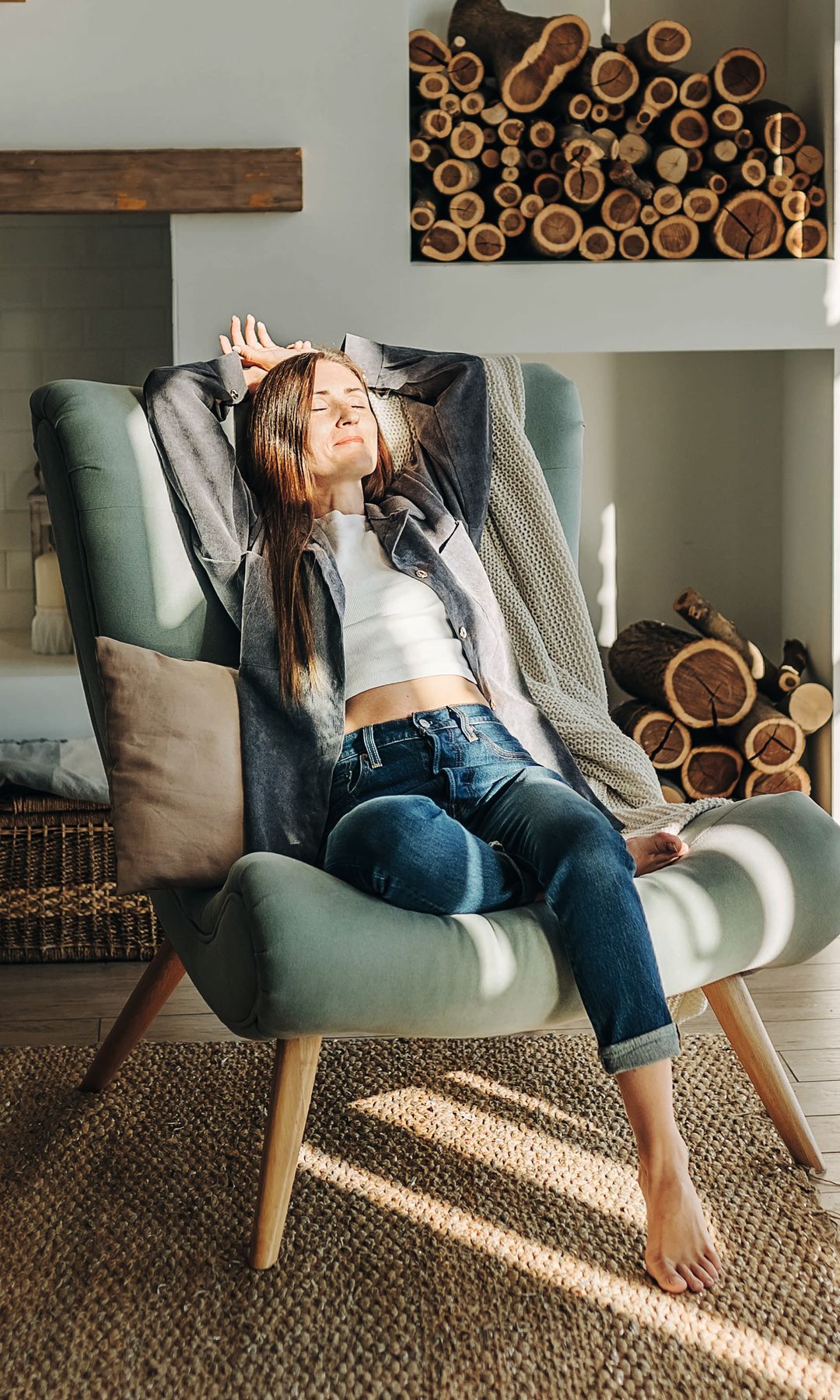 Mujer con jeans y chaqueta gris relajada en sillón verde en sala soleada.