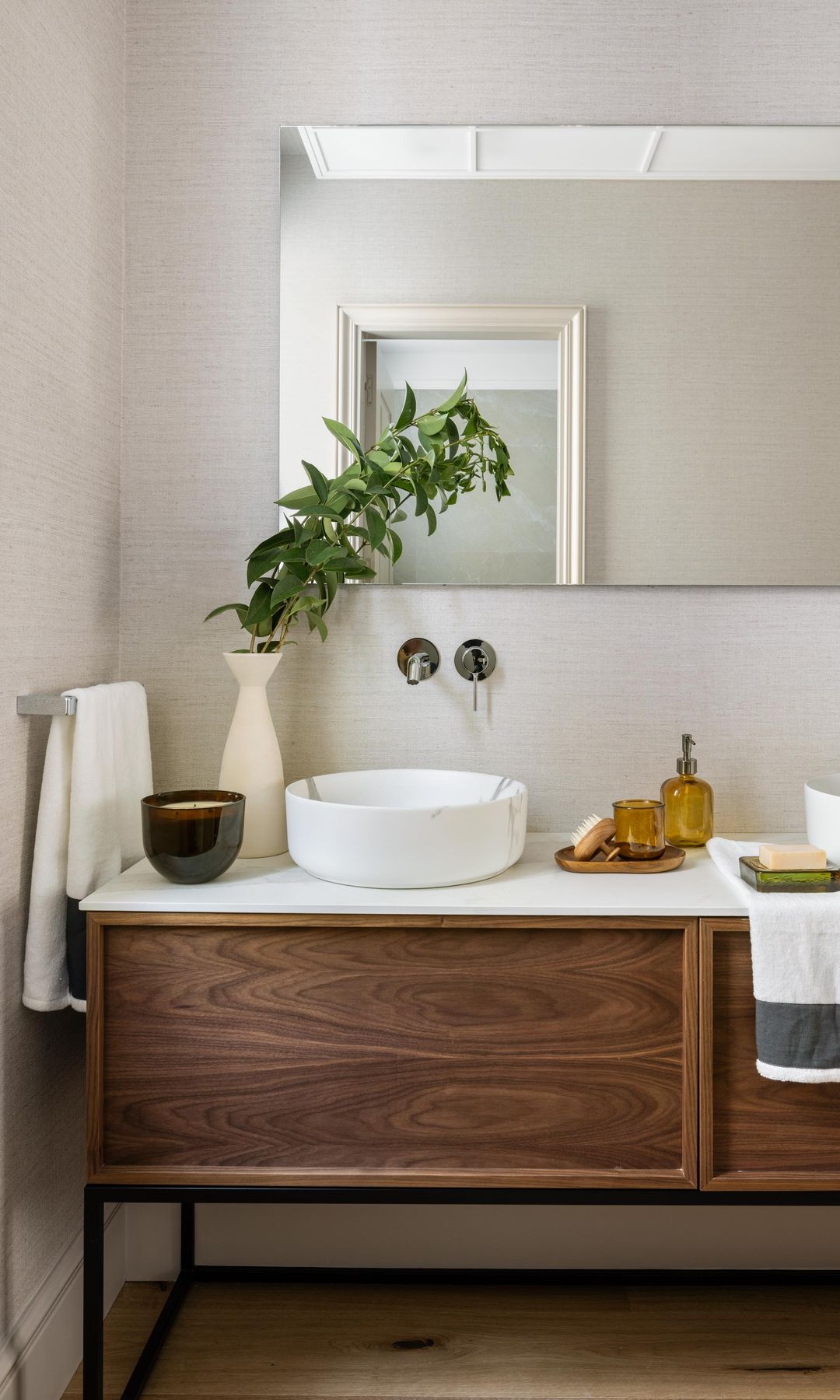 Baño con mueble de lavabo de madera oscura, espejo, velas, bandeja con jabonera, jarrón con rama verde y toallas