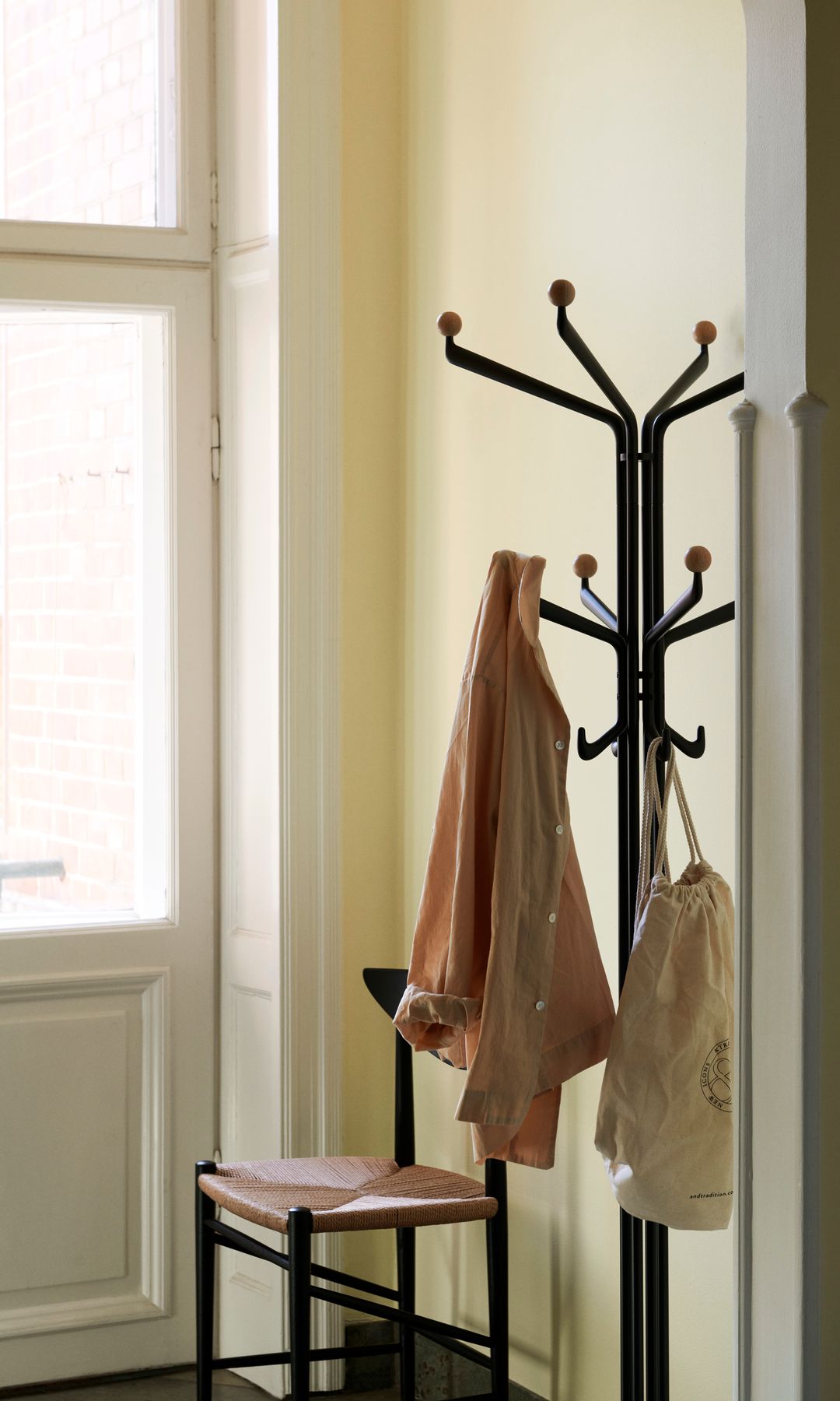 Recibidor con perchero tipo árbol negro con chaqueta y bolsa, silla negra con el asiento de enea, puerta de cristal y madera blanca