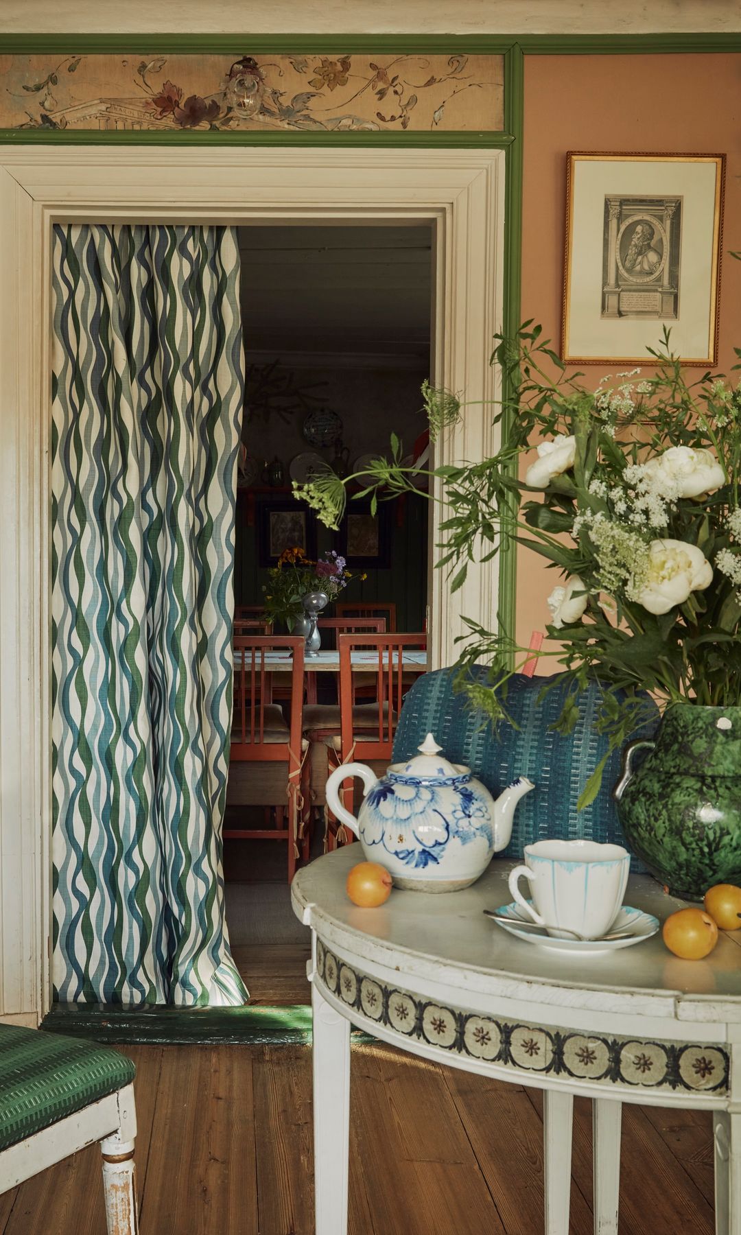 Comedor con cortina separando la cocina, mesa redonda blanca, silla tapizada en verde, jarrón verde con flores, tetera, taza, cuadros en la pared