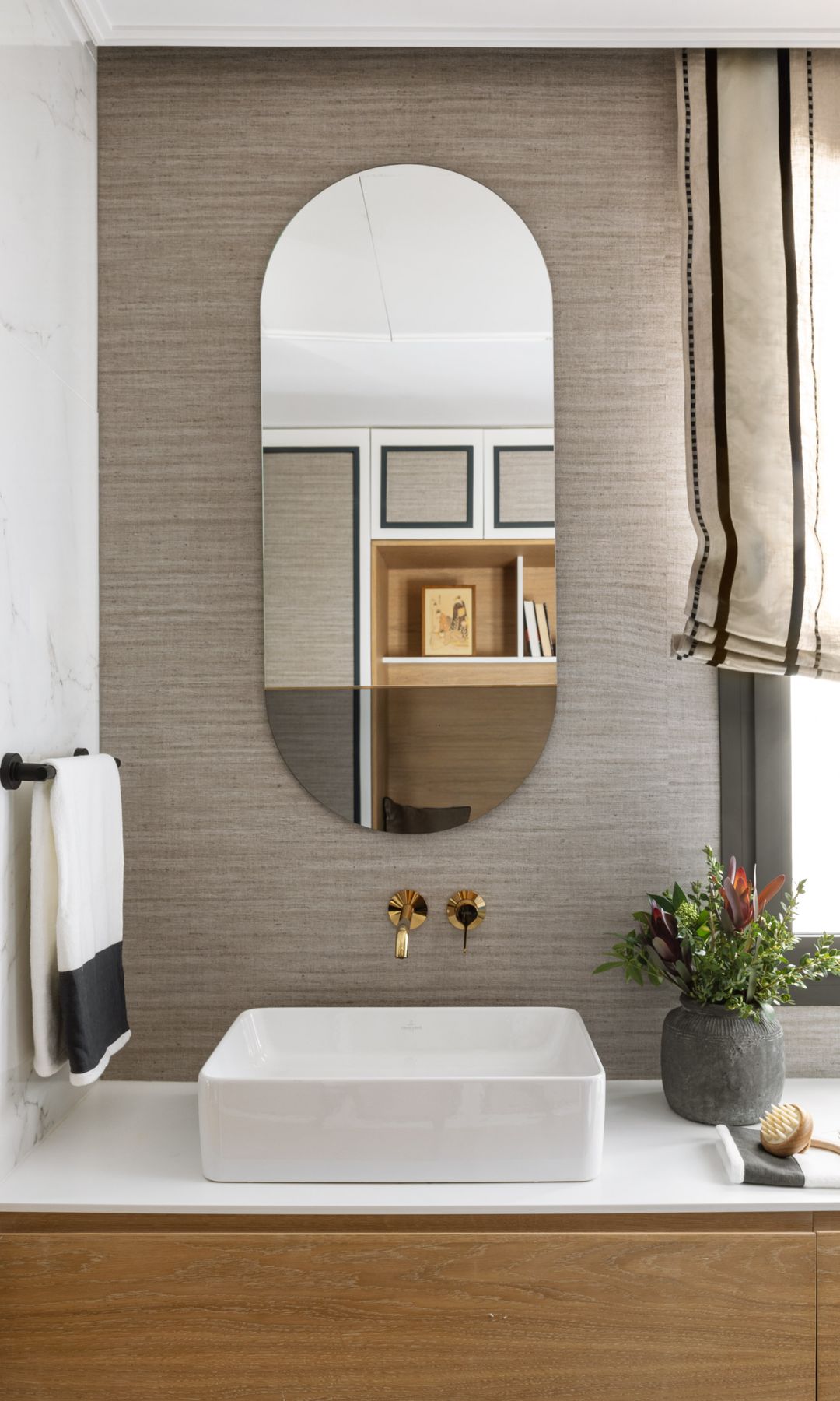 Baño con papel pintado, estor, lavabo cuadrado, espejo ovalado, jarrón con flores y grifería dorada 