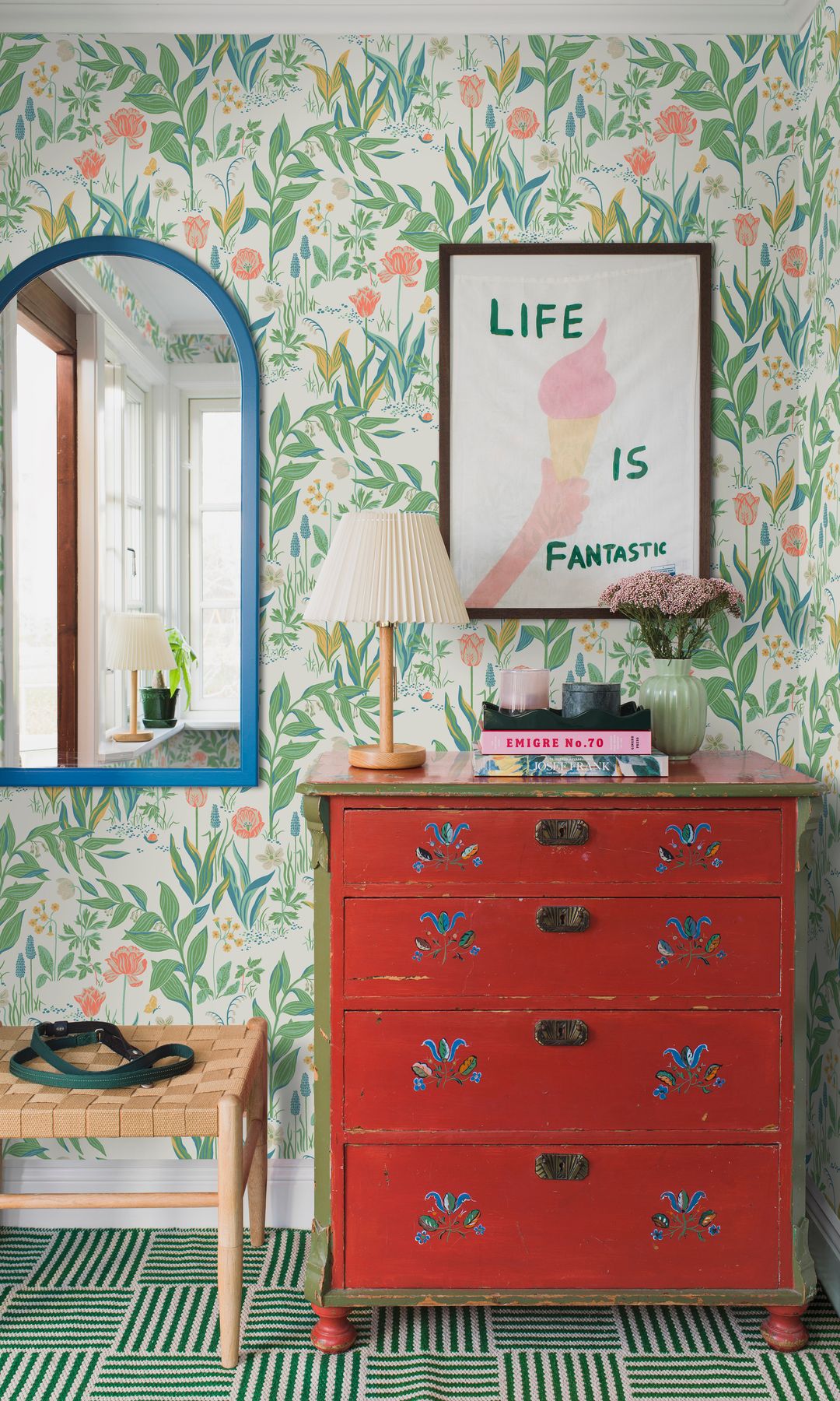 Rincón con cómoda de color rojo, papel pintado de flores, espejo con el marco azul, banqueta de fibras vegetales, cuadro en la pared, lámpara de sobremesa 
