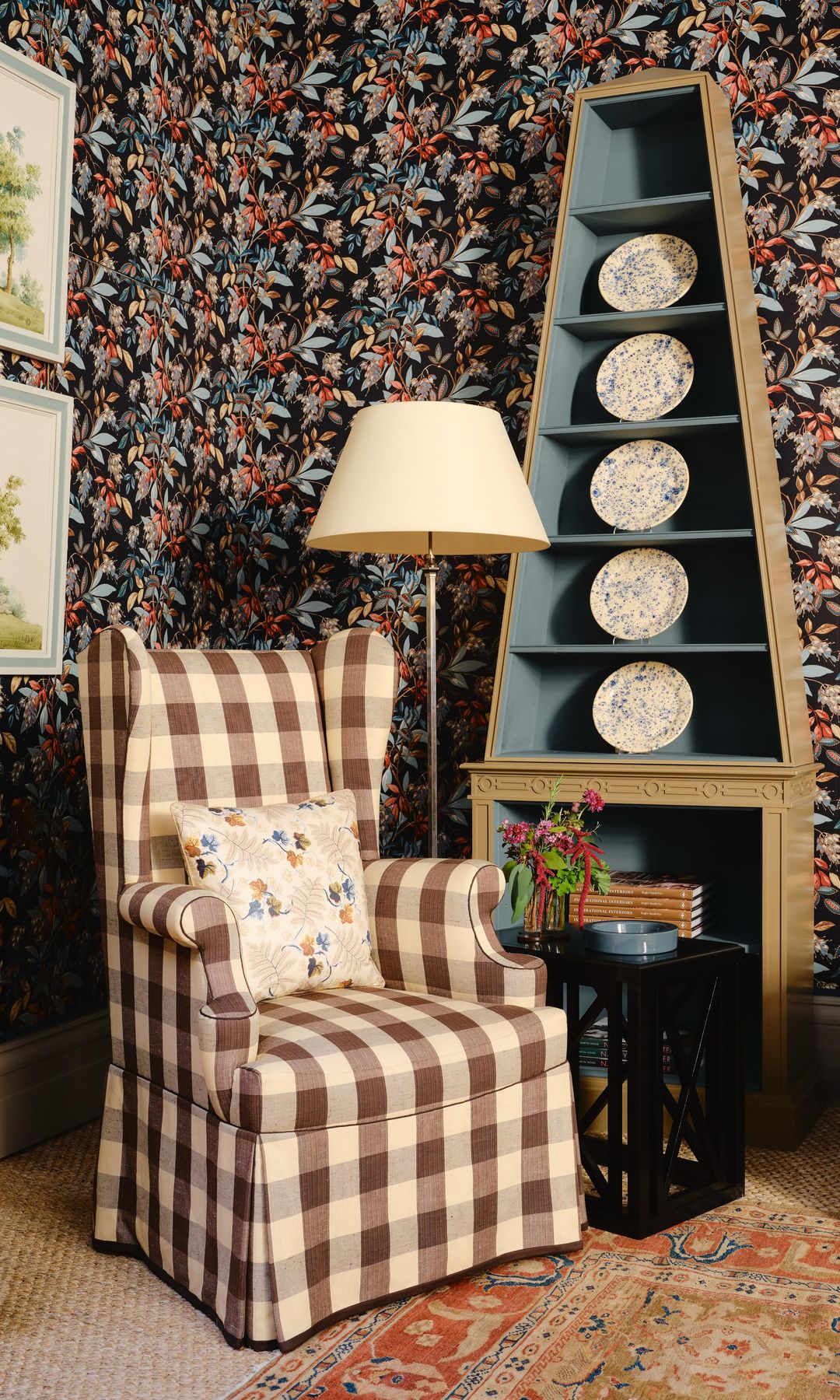 Salón con chimenea con papel pintado de flores negras y rojas, orejero de cuadros marrones y beiges, estantería con platos, alfombra, mesa auxiliar negra