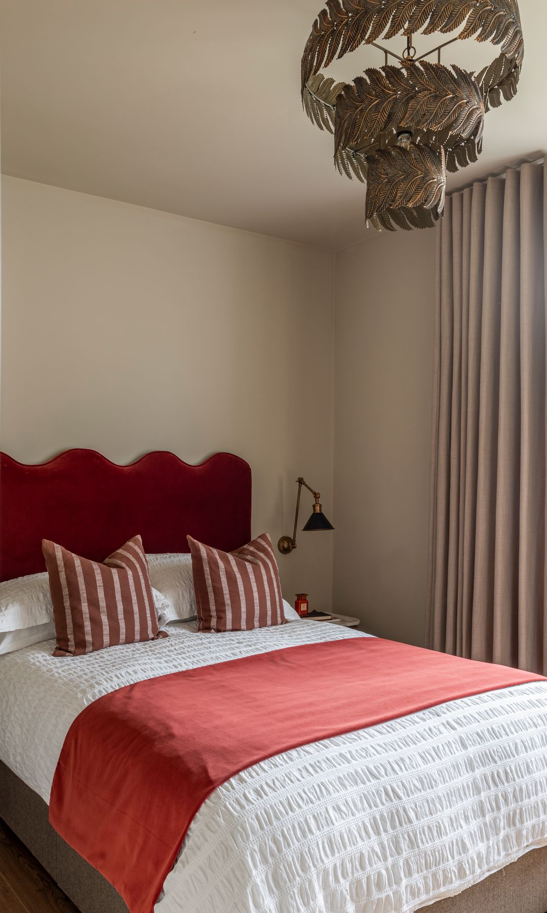 Dormitorio con cabecero tapizado de curvas y en color burdeos, lámpara de techo, paredes arena, ropa de cama blanca, cojines, manta roja
