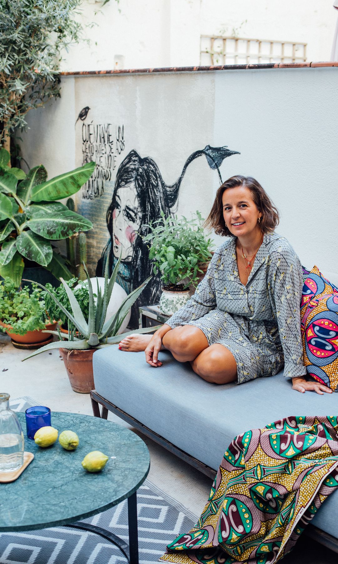 La interiorista Bárbara Aurell sentada en el sofá del patio de su casa de Barcelona, alfombra, mesa redonda de centro, mural pintado en la pared, plantas