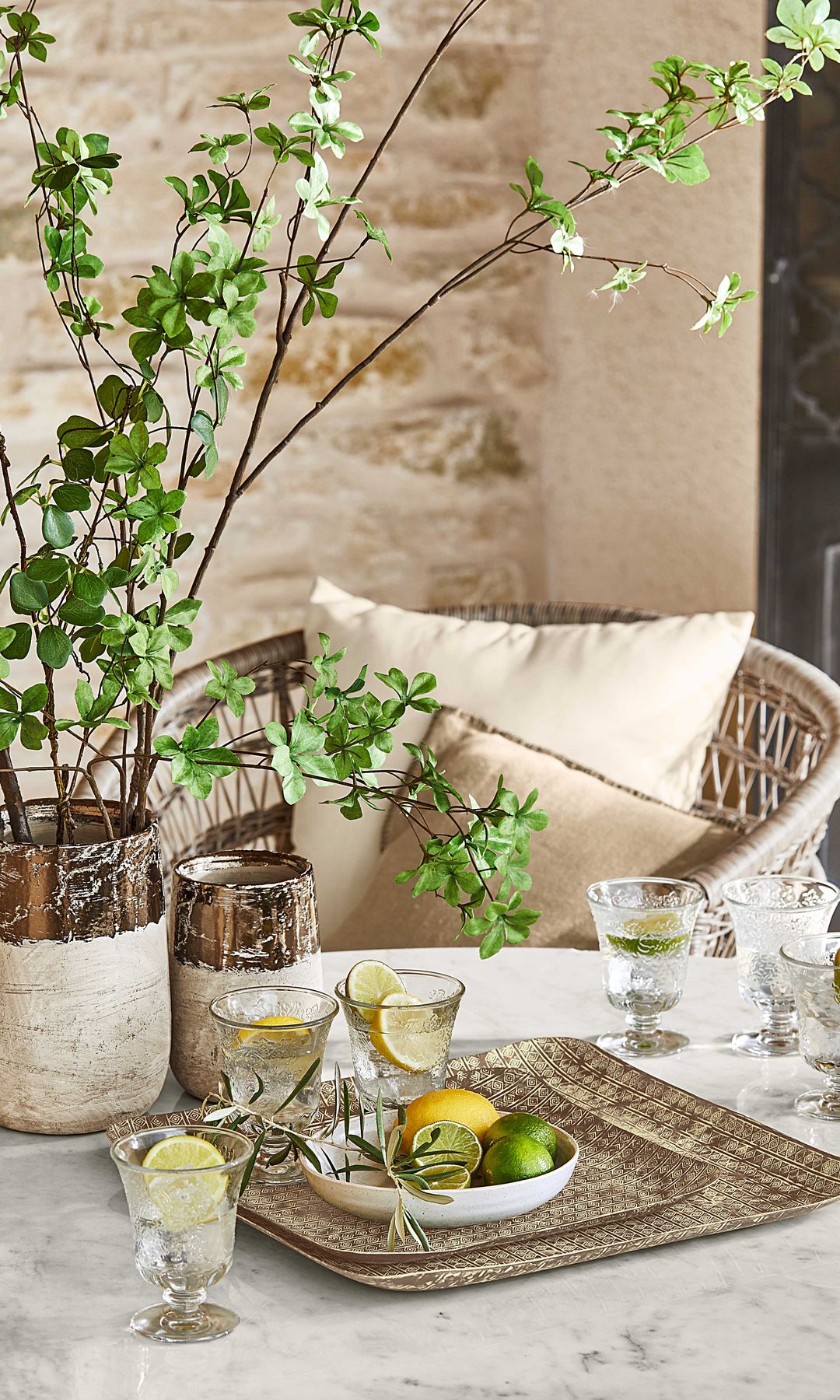 Mesa de jardín con bandejas, jarrón, cristalería, butaca de fibras vegetales y cojines beige
