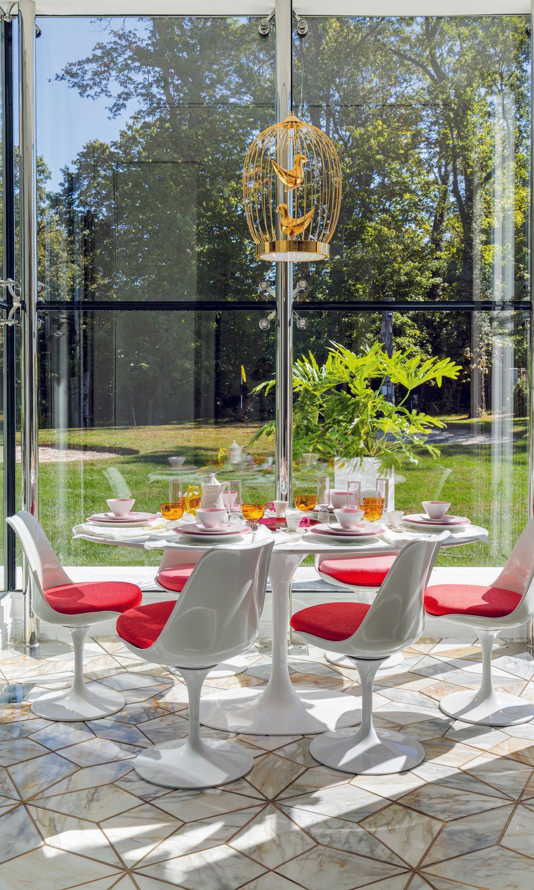 Comedor con mesa redonda y sillas blancas con cojines rojos. Pajarera dorada con dos pájaros dorados colgando del techo. Vista a un jardín con césped y árboles.