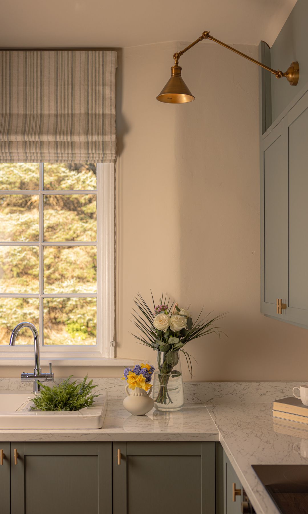 Cocina pequeña con ventana, estor de rayas, muebles grises, aplique de pared dorado, encimera de mármol, jarrón con flores 