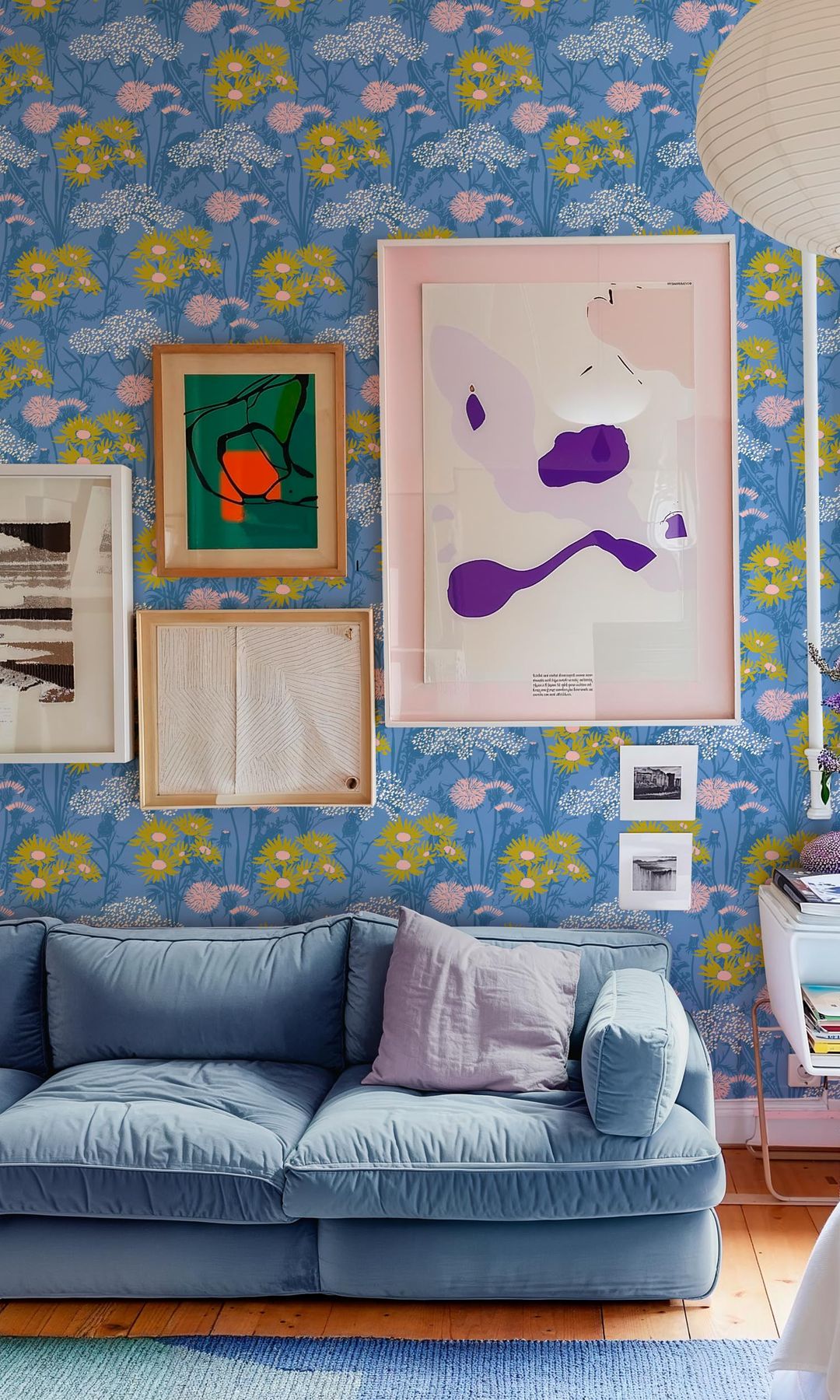 Salón con papel pintado de flores azul y rosa, cuadros en la pared, sofá de terciopelo azul claro, mesa con libros, alfombra azul, lámpara de techo de papel