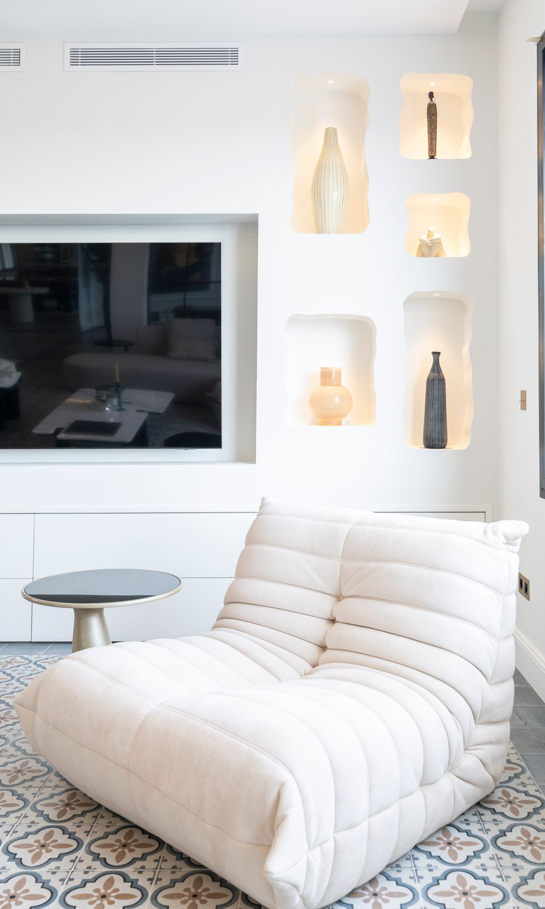 Rincón de lectura con sillón blanco, suelo hidráulico, mesa auxiliar y mueble a medida con la tele colgada y hornacinas 