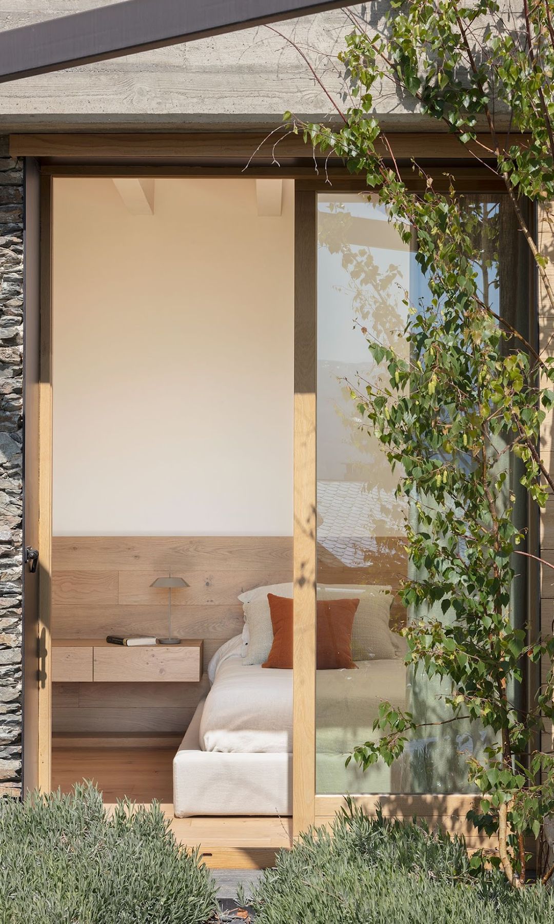 Vista del dormitorio desde el jardín, con cabecero de madera con mesita de noche integrada, ropa de cama blanca, cojín marrón 
