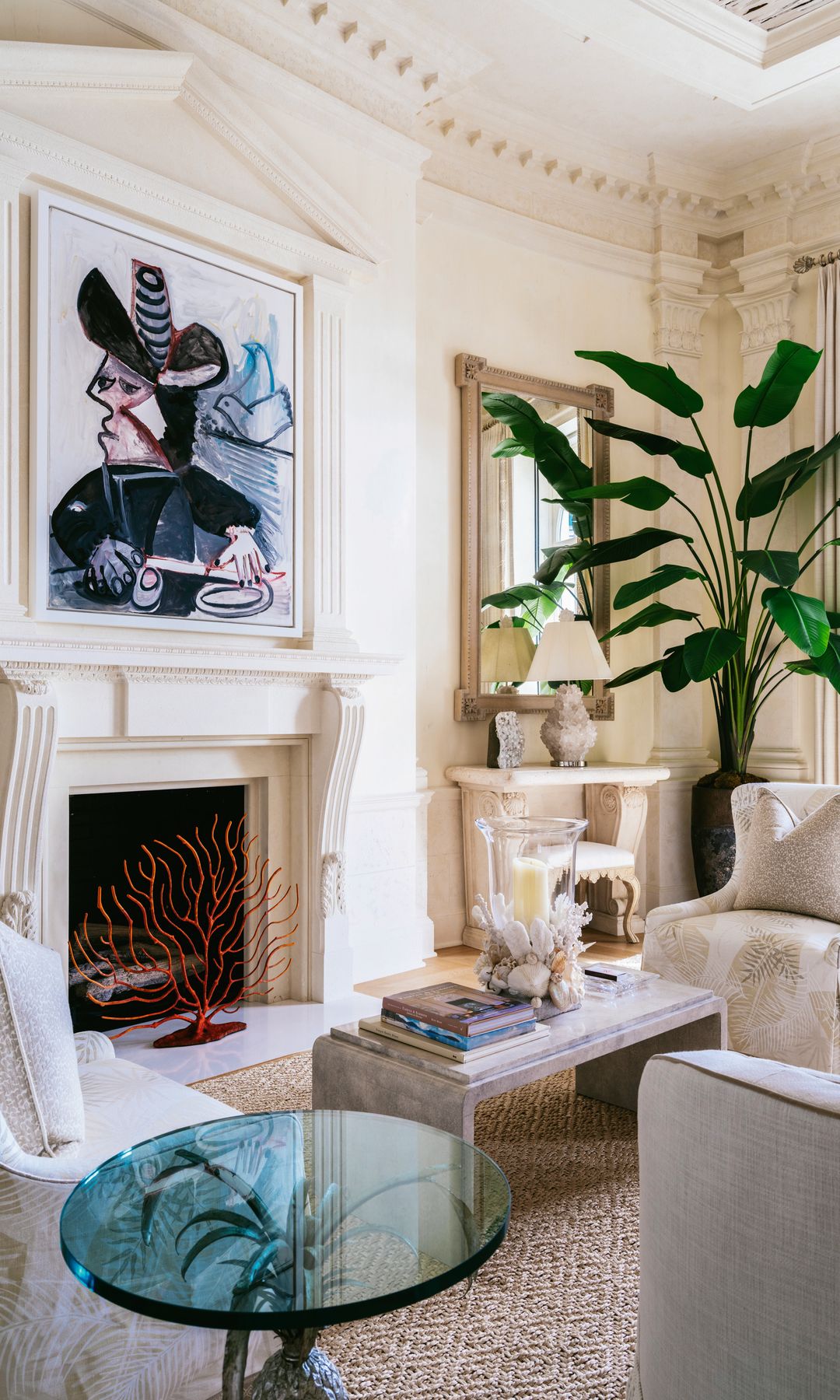 Un cuadro de Picasso "Mousquetaire aux oiseaux" sobre una chimenea en una sala blanca con una mesilla de cristal azul y una plana al fondo