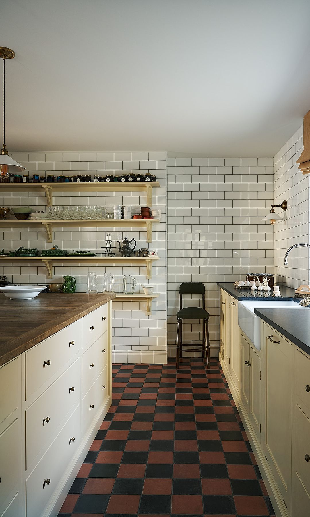 Cocina con muebles clásicos de color crema, azulejos blancos brillantes, tipo metro, suelo de damero marrón y negro, isla con encimera de madera, grifo antiguo