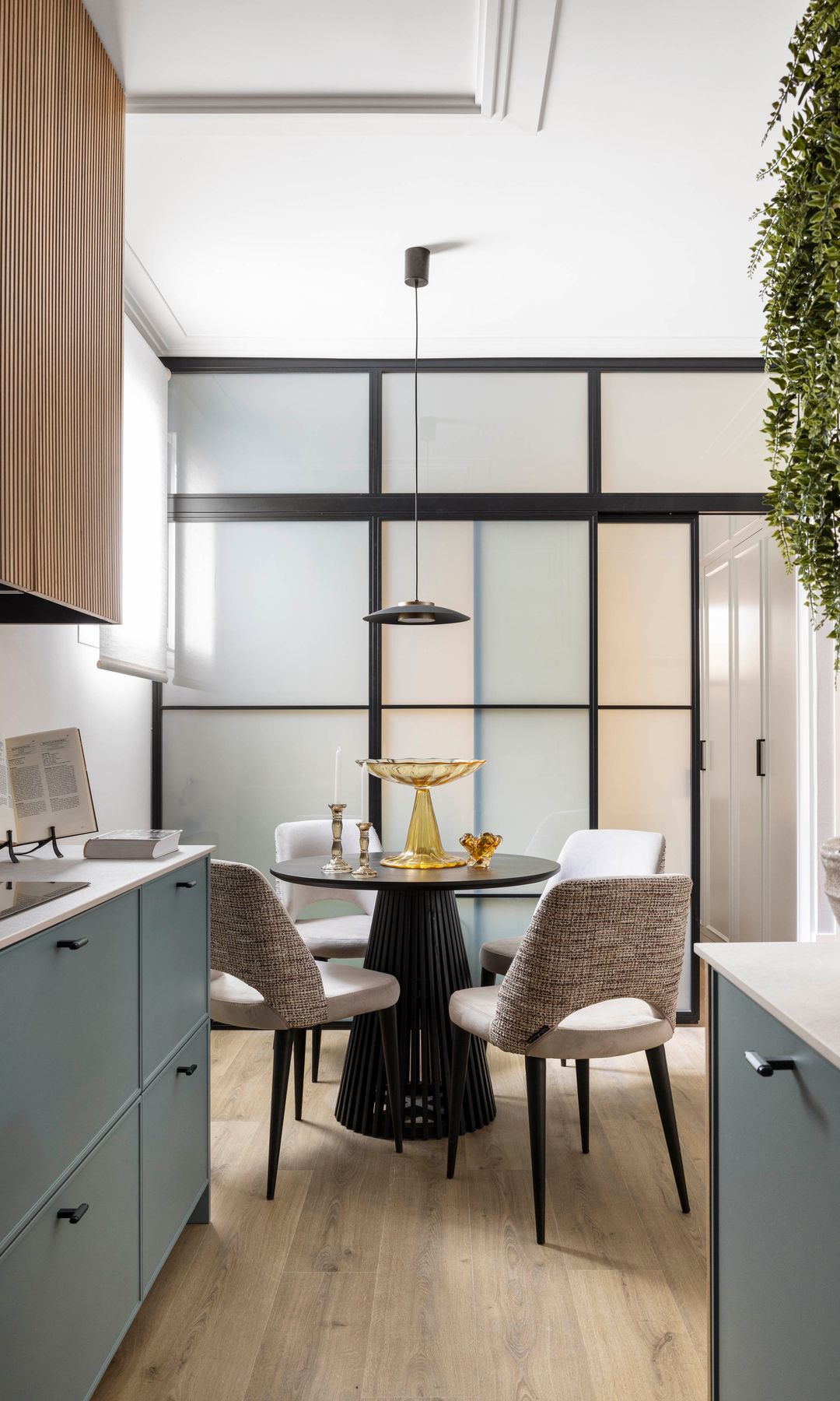 Cocina con comedor con mesa redonda, sillas tapizadas, tabique de cristal con cuarterones negros, muebles de cocina grises, armarios altos madera