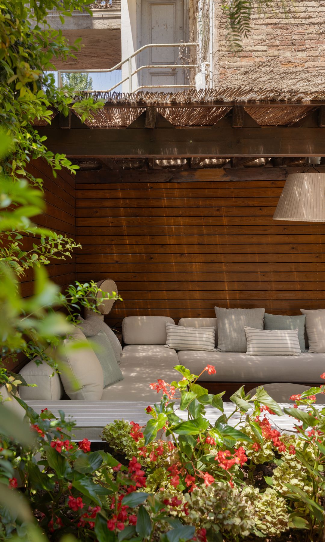 Salón exterior bajo pérgola de madera y caña, sofás grises, cojines, plantas con flores y lámpara de techo blanca