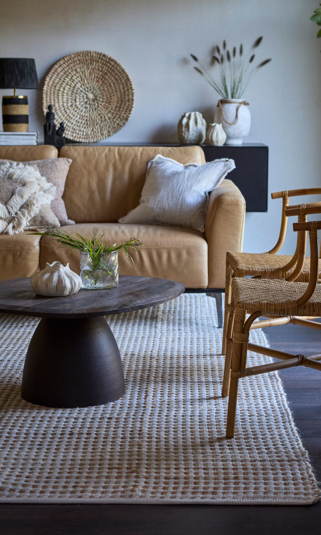 Salón con sofá de cuero marrón claro, alfombra de fibras vegetales, planta en una maceta, mesa de centro de madera oscura, cojines en el sofá, lámpara de sobremesa, sillas de enea 