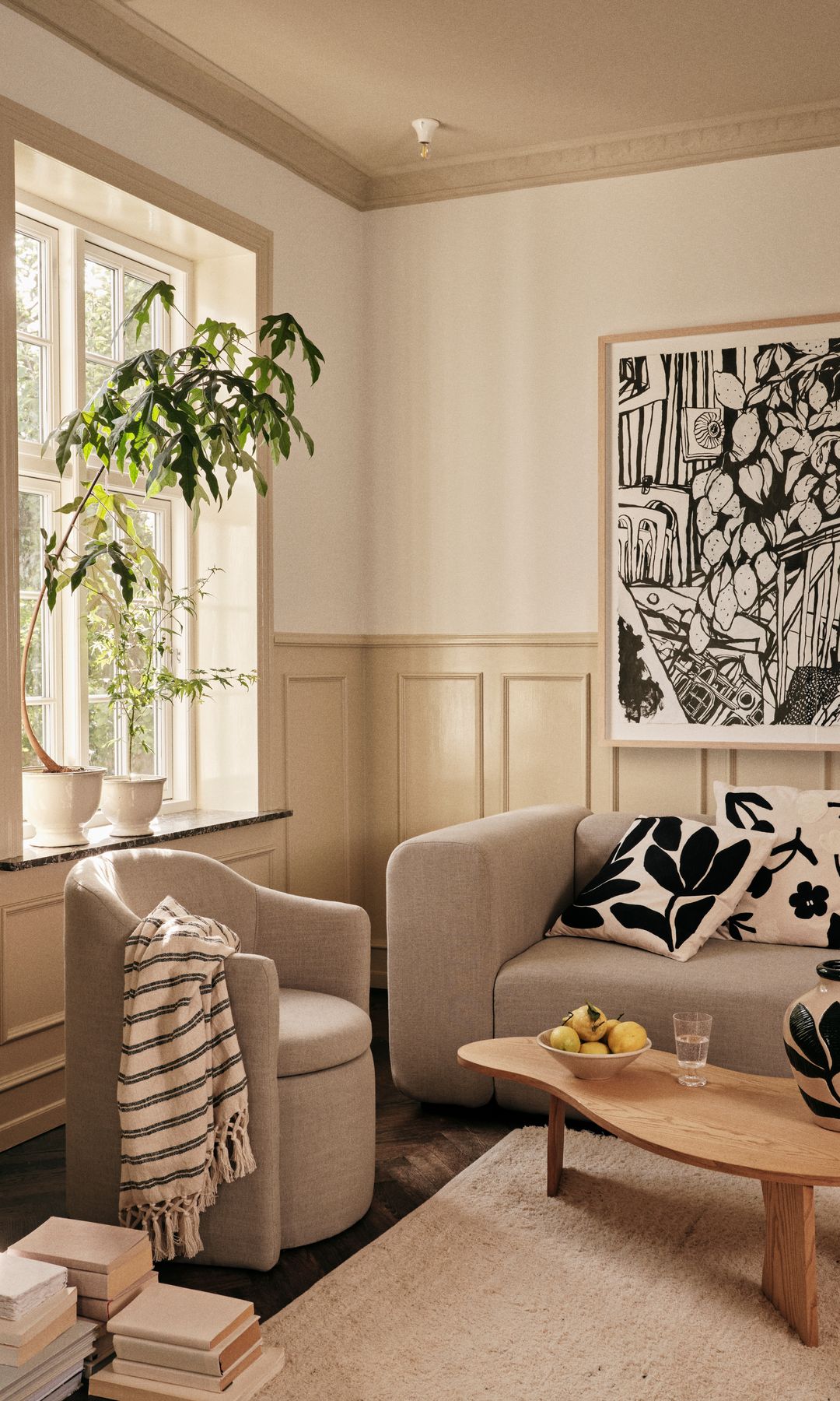 Salón con friso de madera blanca, sofás beiges, alfombra de fibras vegetales, mesa de centro de madera, plantas en el alféizar de la ventana, cuadro en blanco y negro y libros en el suelo