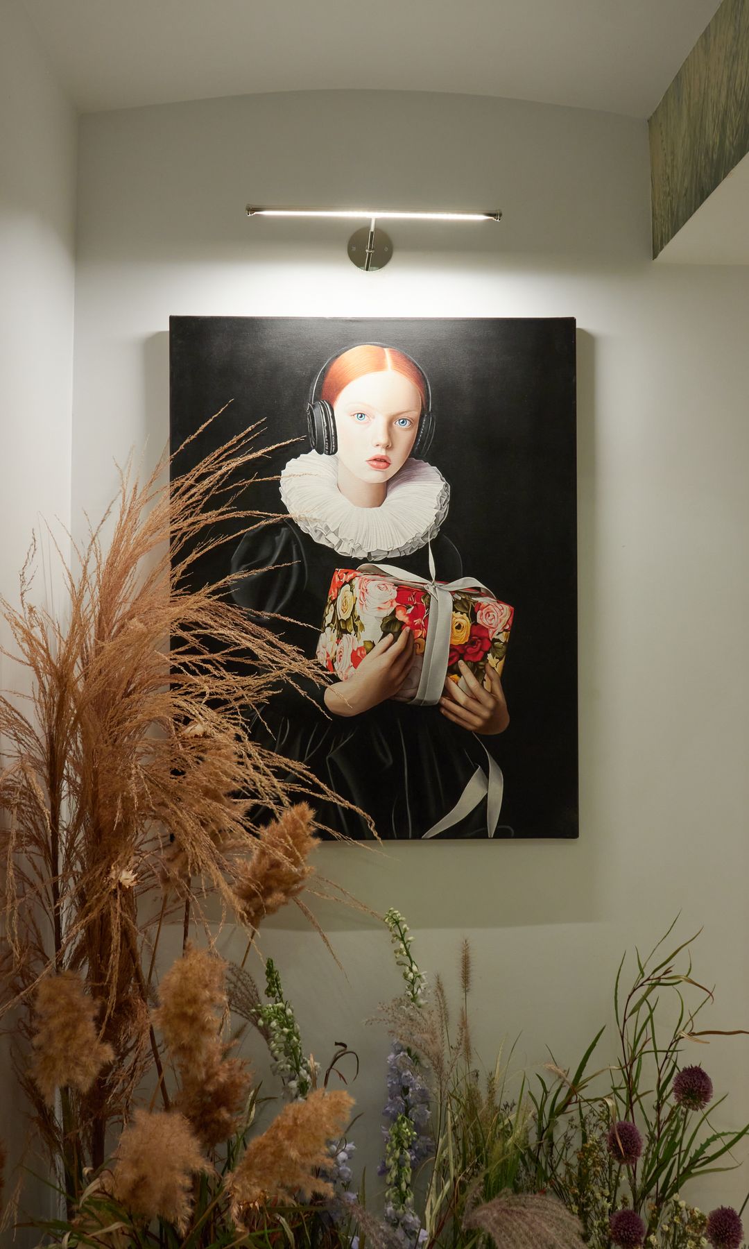 Cuadro colgado en la pared, sin marco con el fondo negro y el retrato de una mujer, aplique dorado sobre él y flores secas en el suelo 