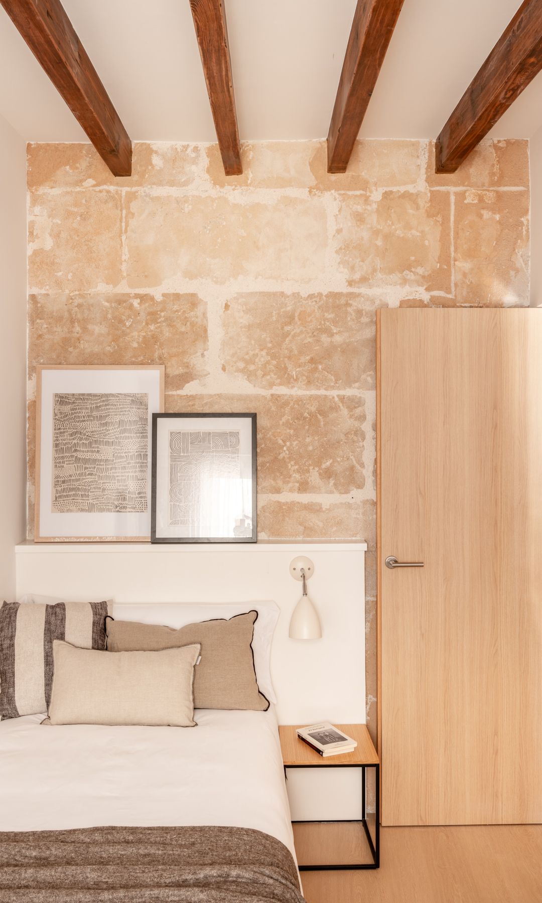 Dormitorio con vigas de madera, cabecero de obra, pared de piedra, puerta de madera, mesita de madera y metálica negra, ropa de cama natural, balda cabecero, cuadros