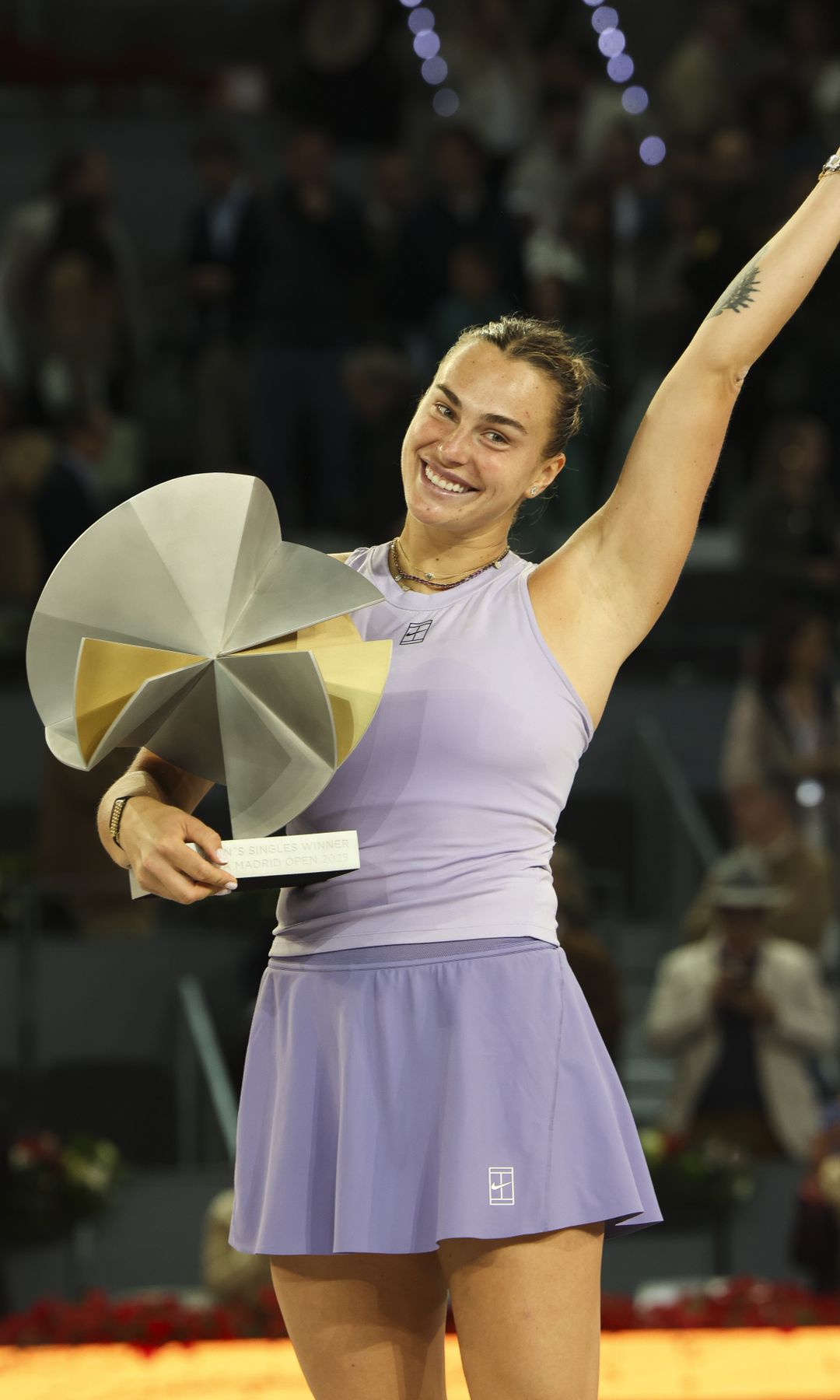 Aryna Sabalenka, con un vestido deportivo morado, sostiene un trofeo y sonríe en una cancha de tenis.