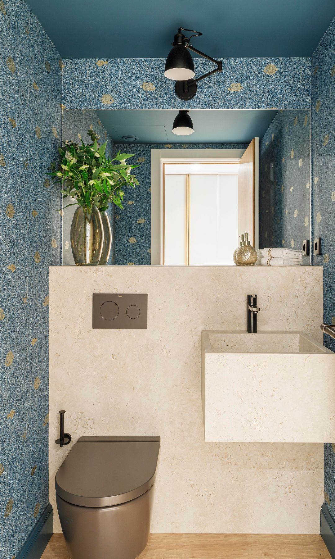 Aseo de cortesía con papel pintado azul de flores, foco en el techo negro, cisterna empotrada, sanitario de color cobre, lavabo suspendido