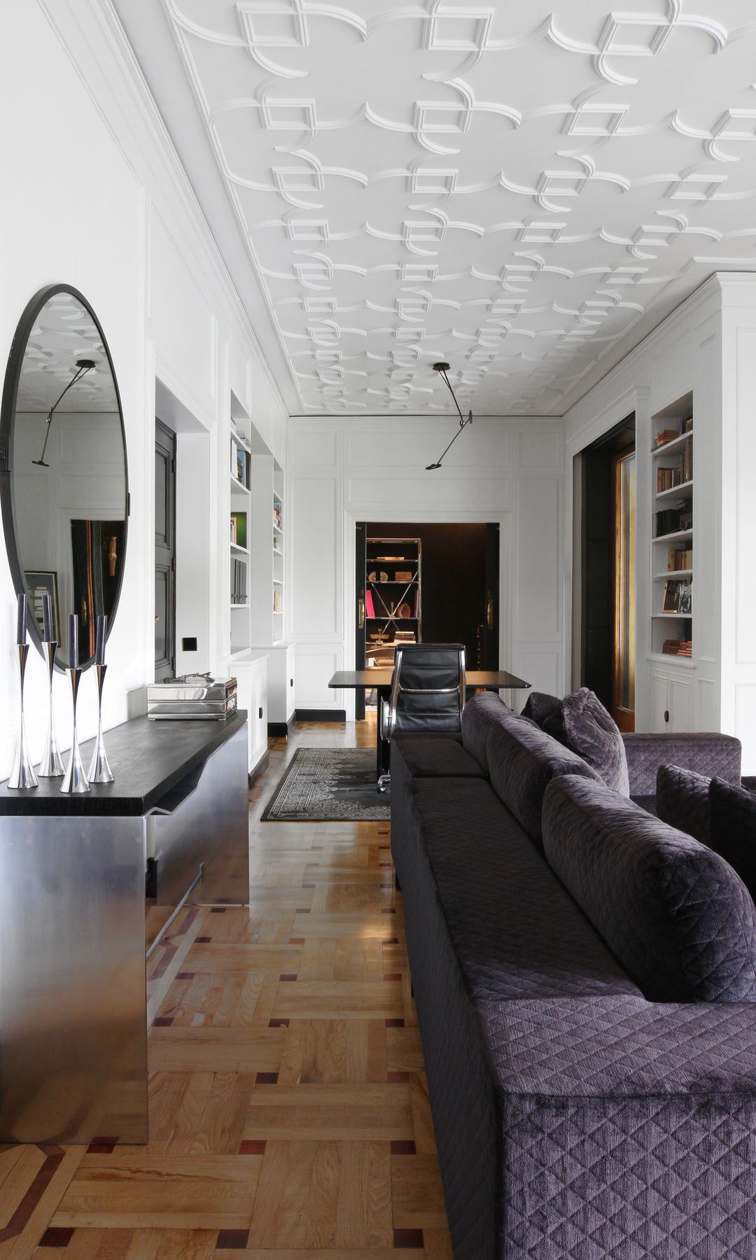 Salón con sofá negro de terciopelo, espejo redondo, consola plateada, despacho, suelo de madera, techo decorado 