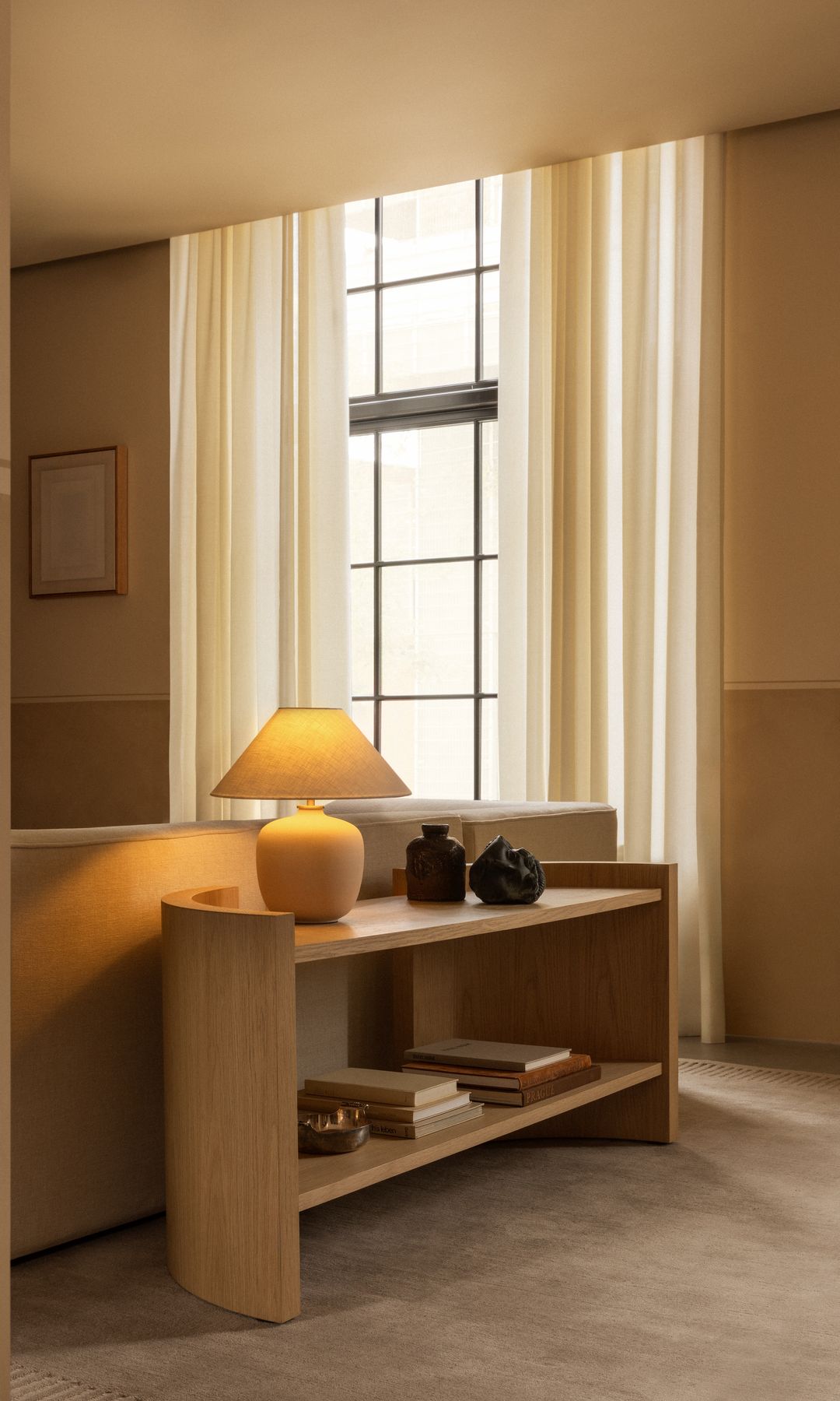 Salón con estantería tras el sofá a modo tabique, lámpara de sobremesa encendida y cortinas beige