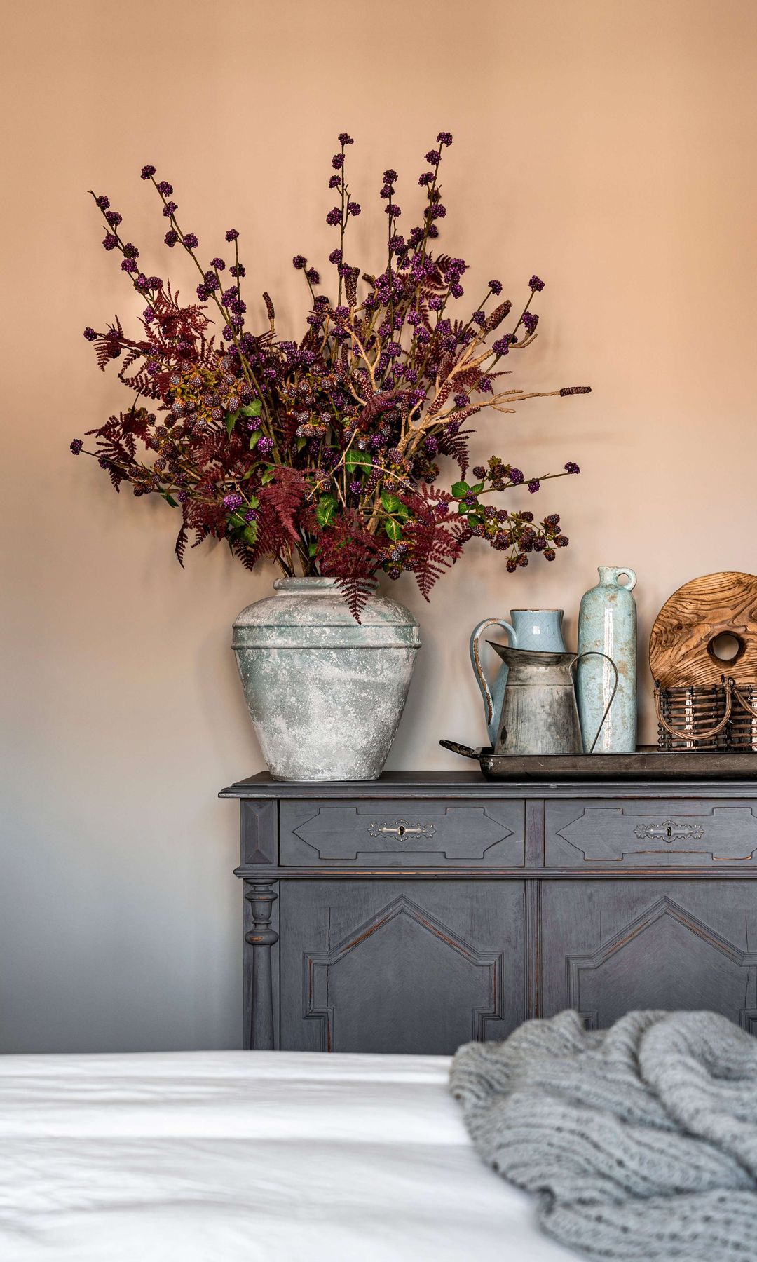 Dormitorio con. aparador de color gris, jarrones de con arreglos florales, paredes de color beige, manta de lana gris sobre la cama 