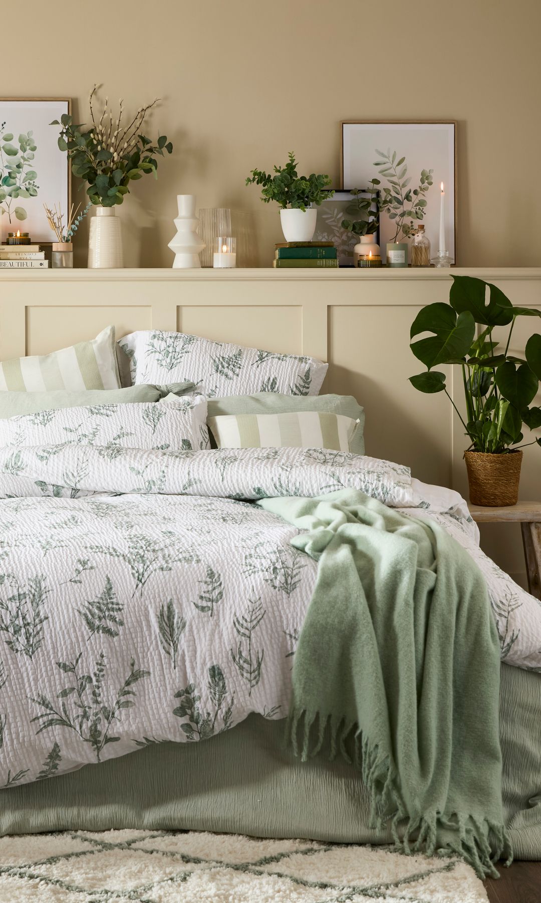 Dormitorio con cabecero de madera beige con balda, plantas, cuadros, funda con motivos de hojas y manta verde, alfombra