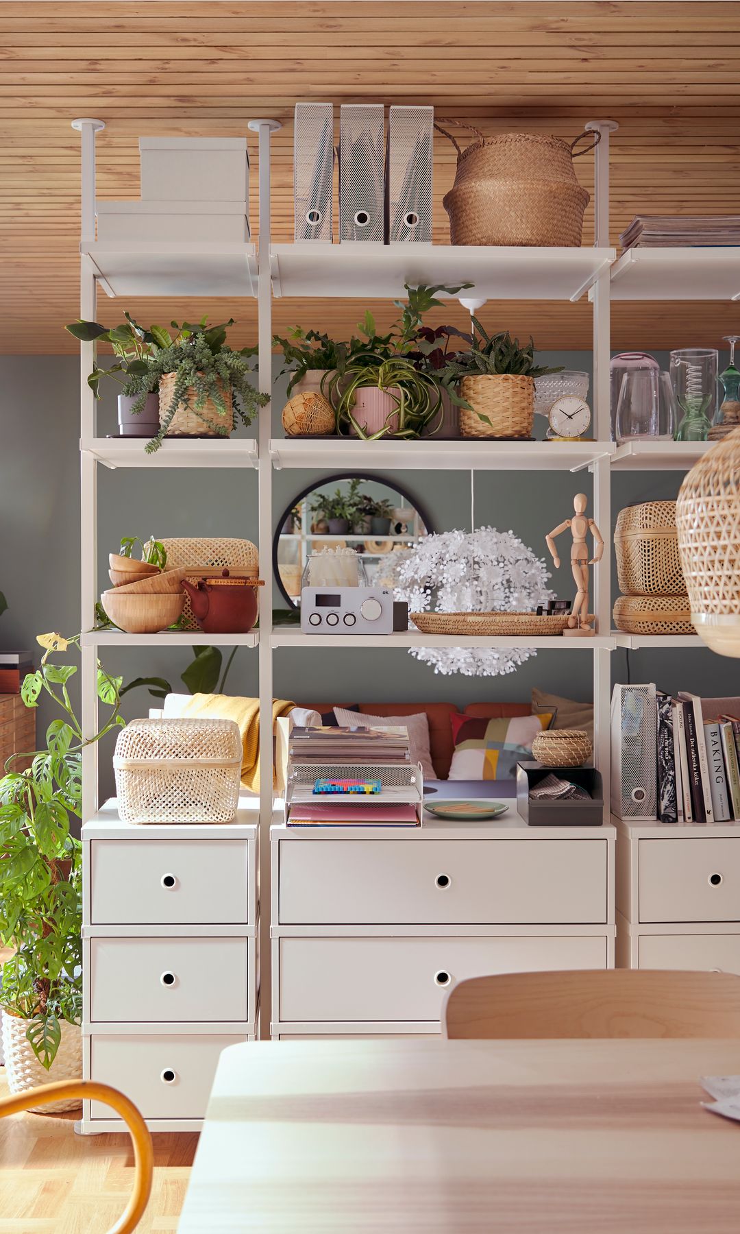 Estantería de IKEA blanca, con zona inferior con cajones y superior con baldas hasta el techo, techo de madera, cestos, macetas, plantas. ventanal, cortinas