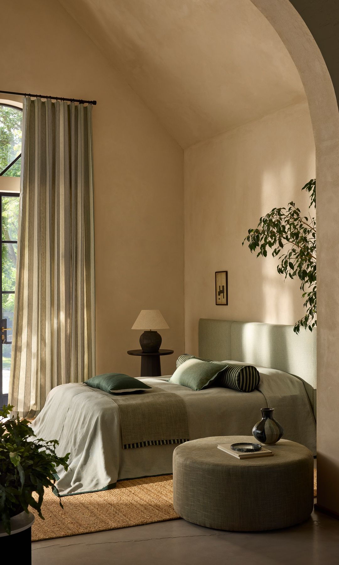 Dormitorio con paredes en beiges, salida al jardín, cabecero tapizado en color verde, ropa de cama verde, cojines, puf, planta, arco en lugar de puerta