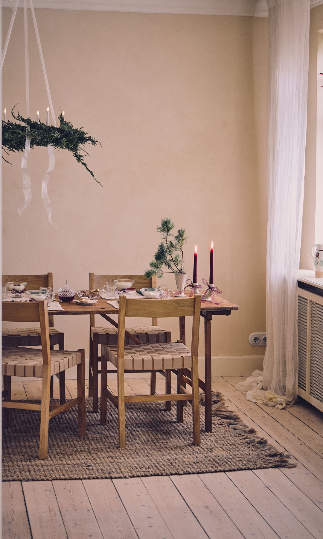 Mesa de comedor vestida de Navidad, con velas, candelabros y arreglo floral en el techo, alfombra de fibras vegetales, cortinas blancas, suelo de madera