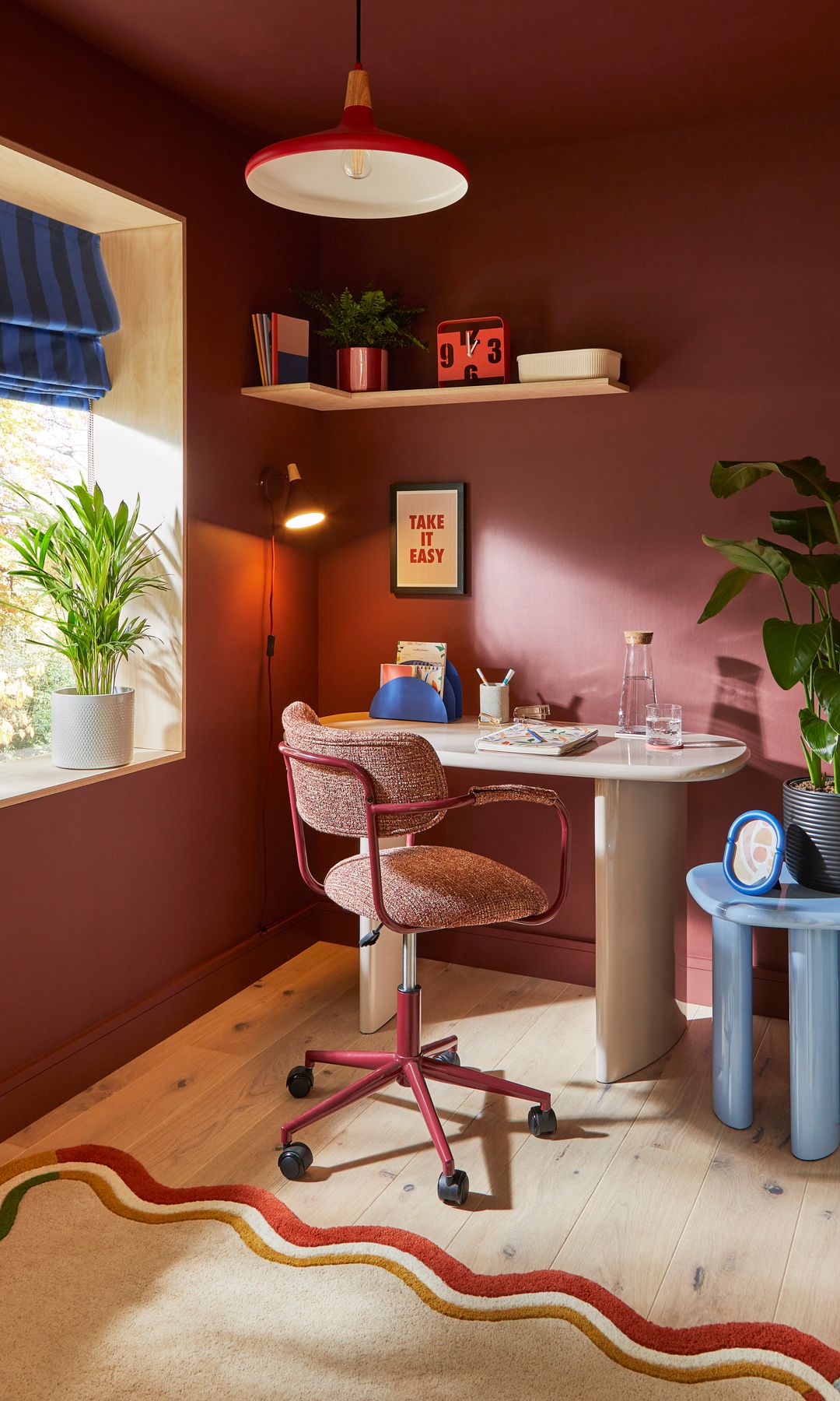 Despacho con paredes en color caldero, lámpara de techo roja, aplique de pared, tipo flexo, silla de oficina con ruedas, balda, estantería con libros y adornos, planta en el alféizar ventana, estor de rayas 