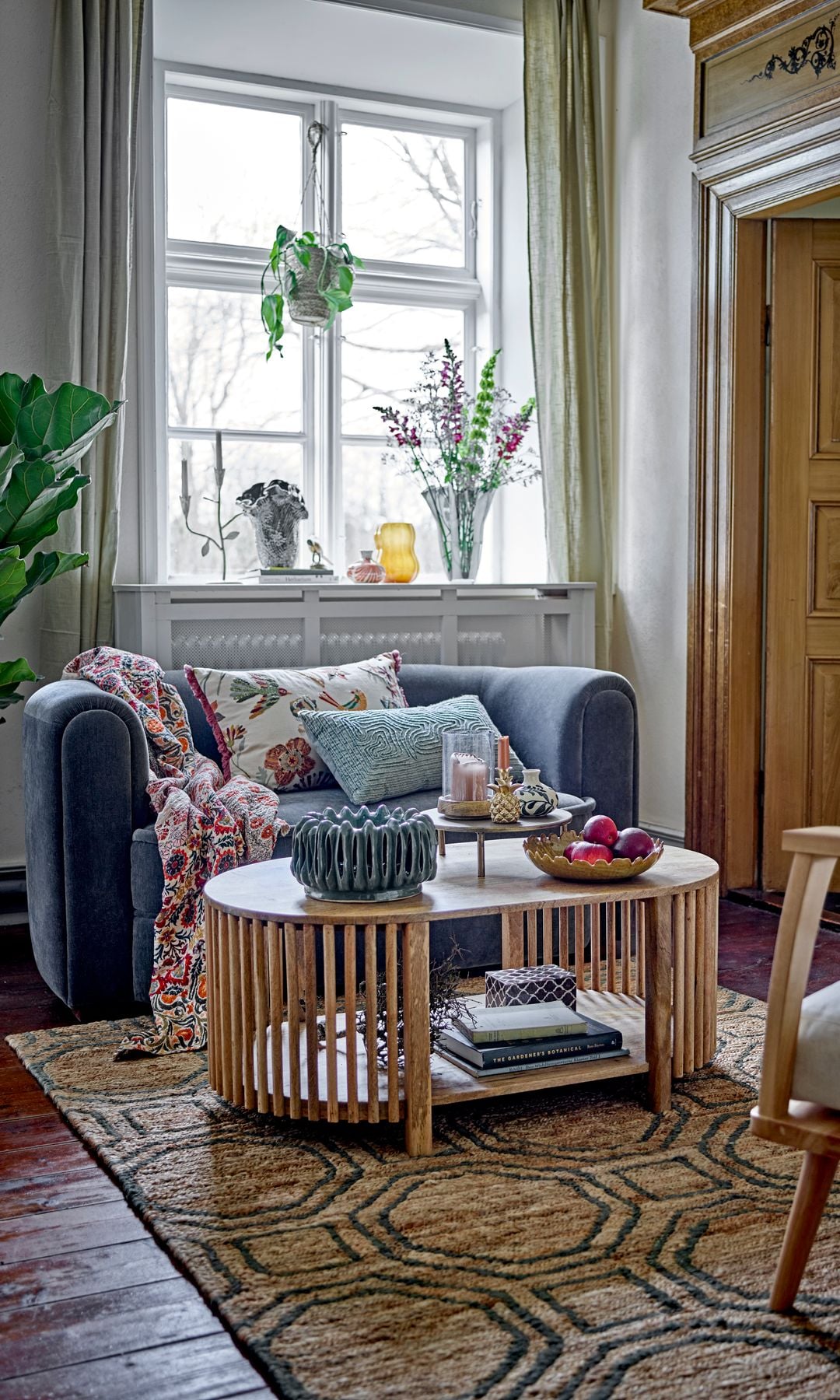 Salón con sofá azul, mesa de centro de listones de madera con balda inferior con libros, adornos sobre la mesa, velas, cojines, alfombra y ventana con plantas colgantes y jarrón con flores