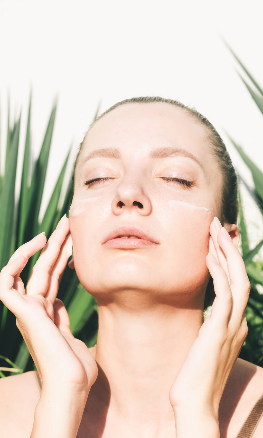 Mujer echándose crema en la cara mientras le da el sol de lleno, con los ojos cerrados mirando hacia arriba.