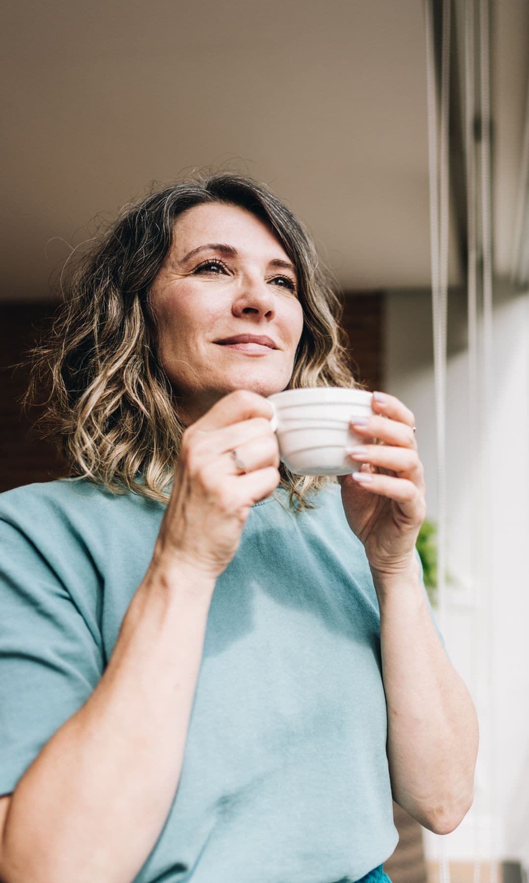 Mujer de 50 años sonriendo con una taza frente a la ventana