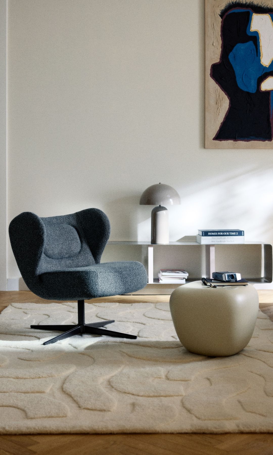 Rincón del salón con butaca de formas curvas en color gris, alfombra, mesa de sobremesa, mesa auxiliar, cuadro pared