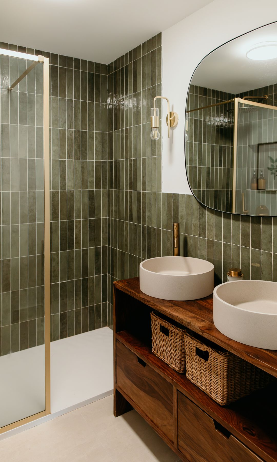 Baño con paredes de azulejos verdes, espejo, mueble de lavabo de madera con la parte inferior abierta con cestos, lavabos sobre encimera redondos 