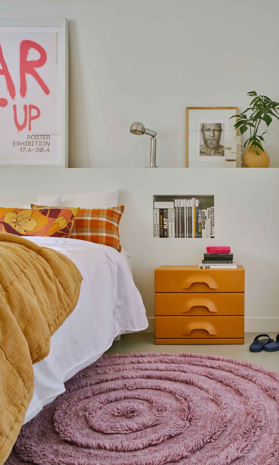 Dormitorio con cabecero de obra con balda, fotografías y cuadro sobre ella, lámpara de sobremesa, mesita de noche naranja, ropa de cama mostaza, alfombra risa, hornacina con libros, cojines amarillos y naranjas 