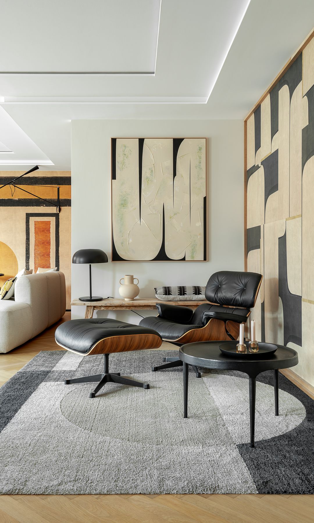 Rincón del salón con chiles longue de los Eames, alfombra gris con motivos geométricos, panel en la pared, lámpara de sobremesa negra, jarrón, cuadro, mesa auxiliar negra