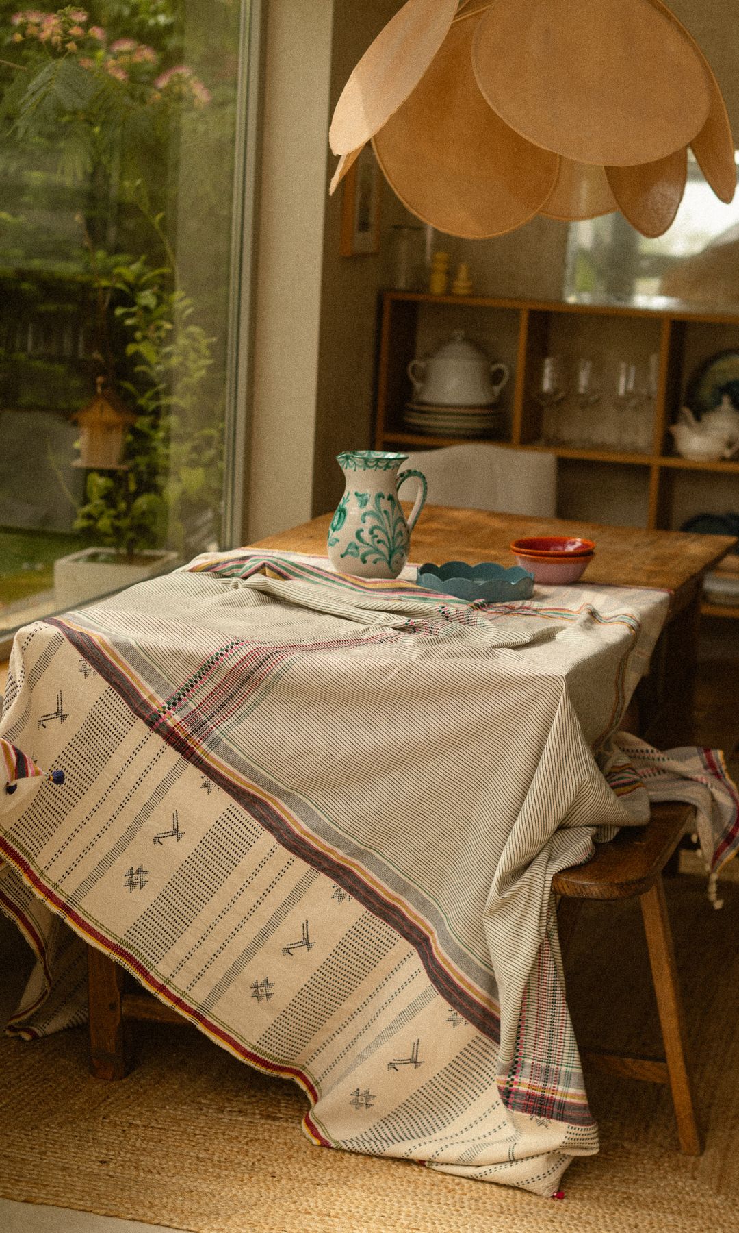 Comedor con mantel con cenefa, jarra de agua de cerámica, platos, lámpara de fibras vegetales, banco