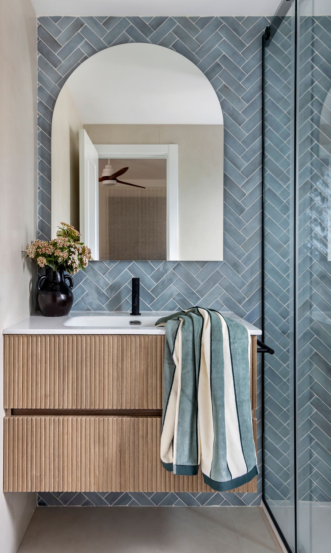 Baño con azulejos azules en espiga, mueble de lavabo de madera, jarrón negro, mampara de ducha con perfiles negros
