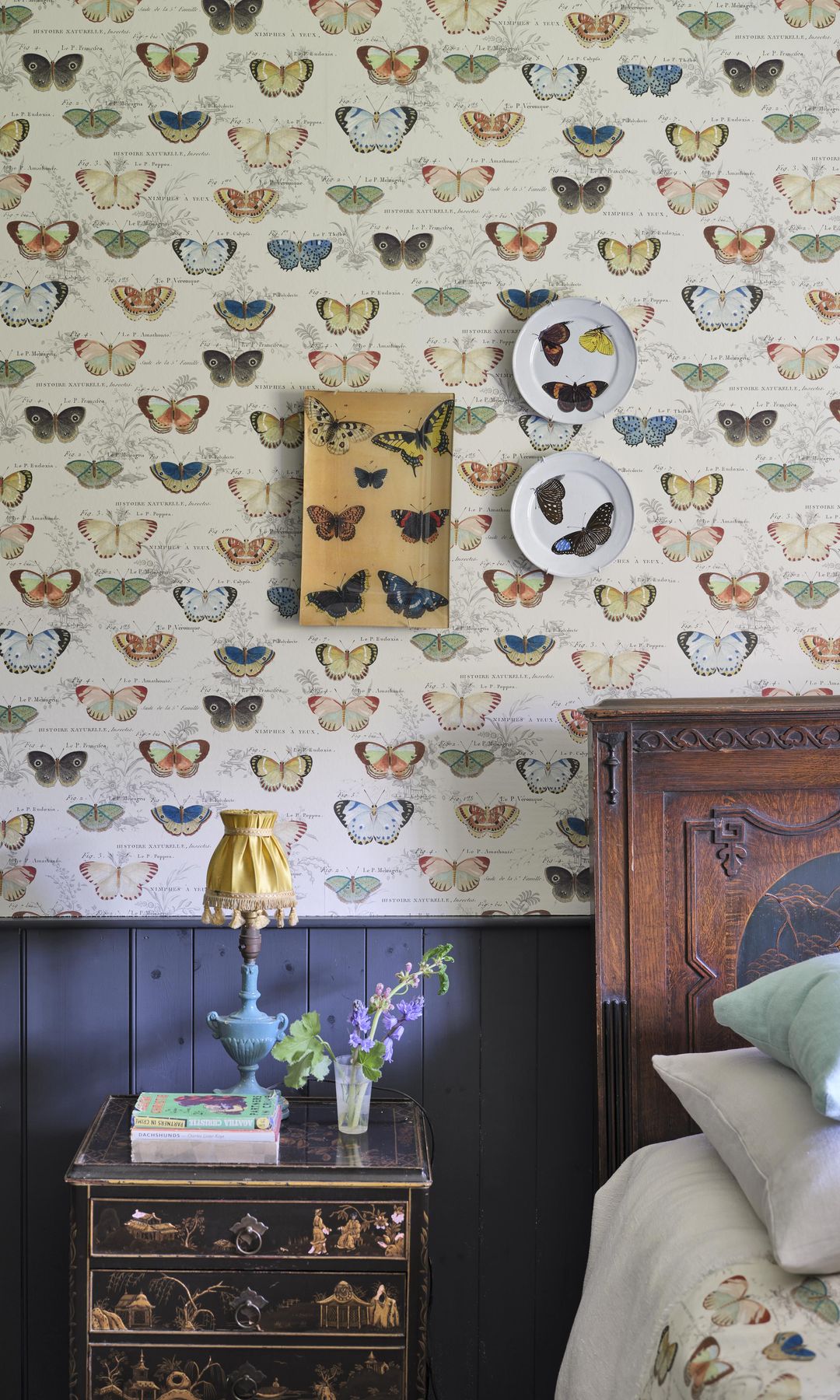 Dormitorio con cabecero de madera oscura, papel pintado en la pared del cabecero, platos en la pared, zócalo de madera pintado de azul, mesita de noche, lámpara de sobremesa azul 