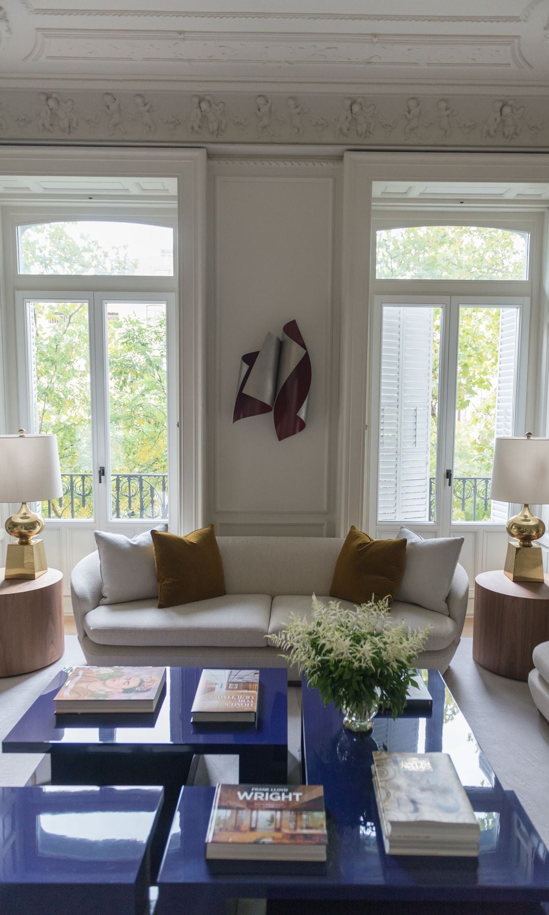 Salón con sofá blanco, mesas de centro azul eléctrico, lámparas de sobremesa, adorno escultura en la pared, ventanales, mesas auxiliares madera  