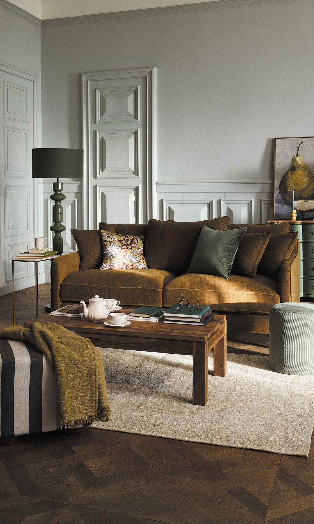 Salón clásico con sofá marrón, suelo de madera, alfombra beige, aparador verde, mesa de madera oscura, puffs, lámpara de pie 