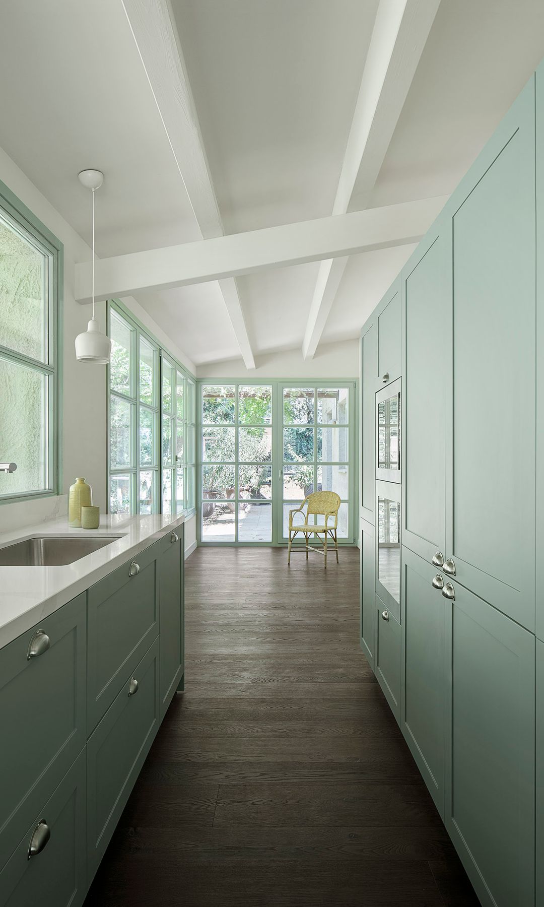Cocina abierta con muebles de color verde claro, con armarios hasta el techo, ventanas y puertas de cristal con el marco del mismo color, suelo de madera oscura
