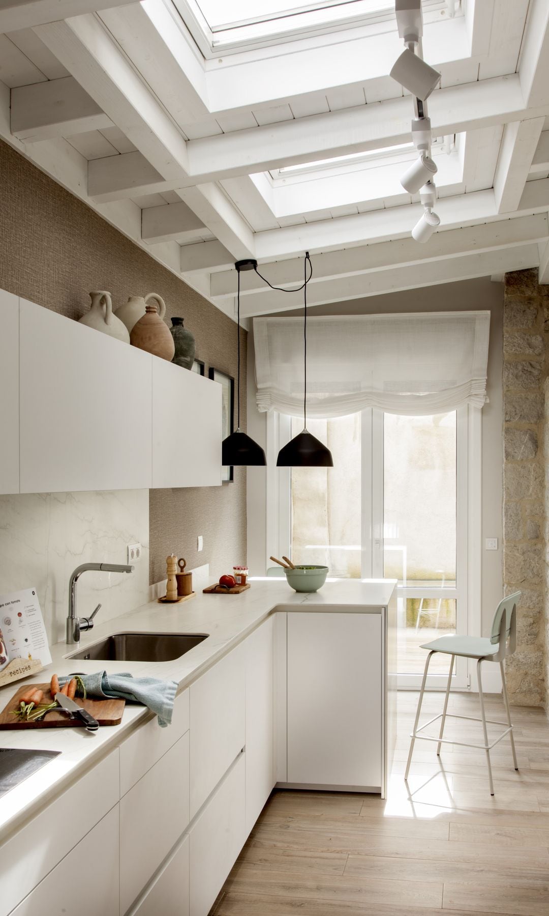 Cocina blanca con ventana de techo Velux, suelo de madera, barra con taburete negro, lámpara de techo negra