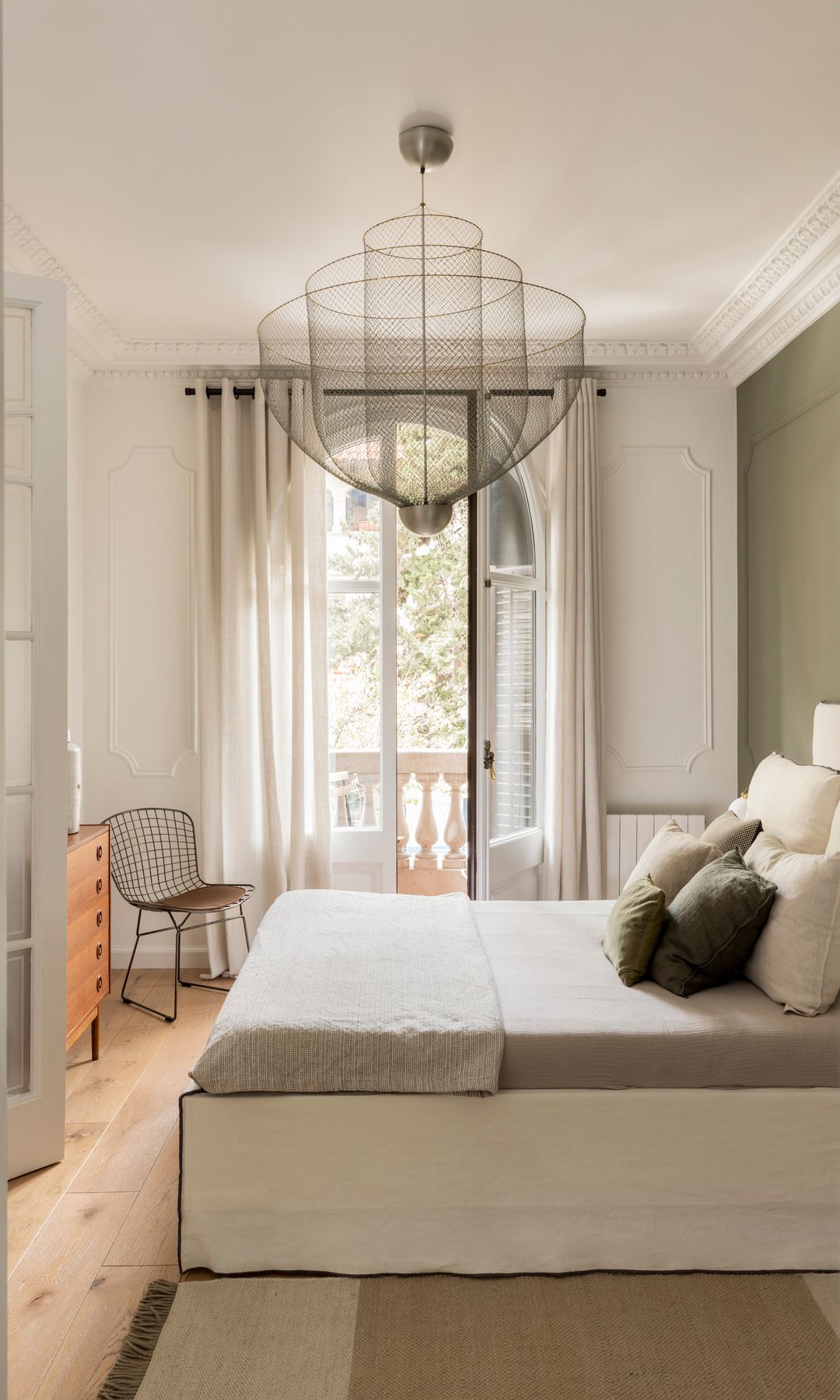 Dormitorio con cama alta vestida en tonos blancos, pared del cabecero verde oliva, lámpara de techo, cortinas blancas, alfombra