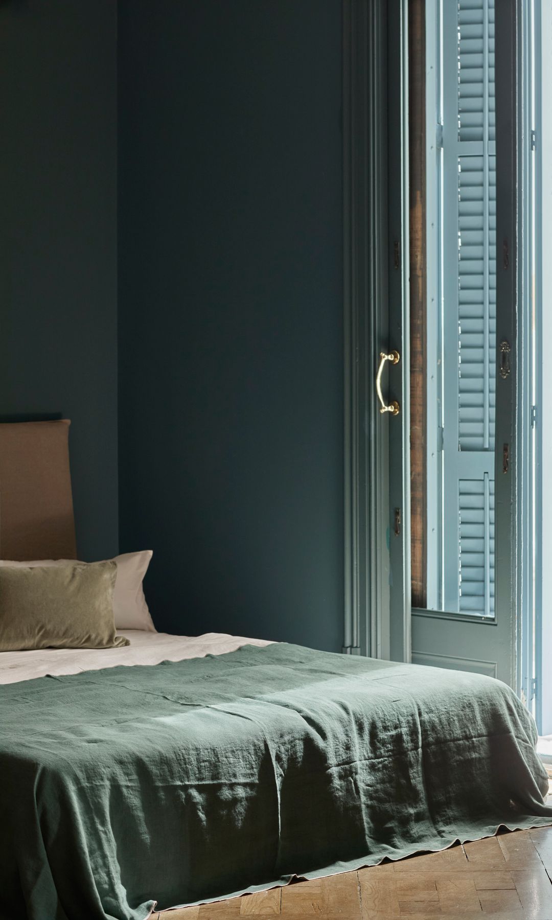 Dormitorio con ventanas abiertas tipo veneciana, suelo de madera, cabecero tapizado, paredes en color azul,. ropa de cama gris